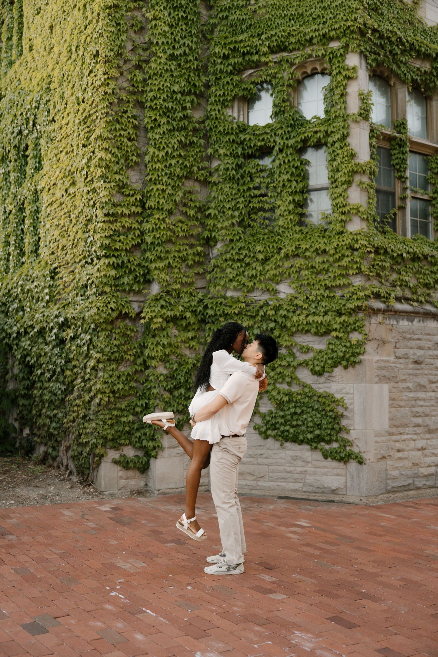 Man lifting woman in front of an ivy-covered stone wall on a brick pathway.
