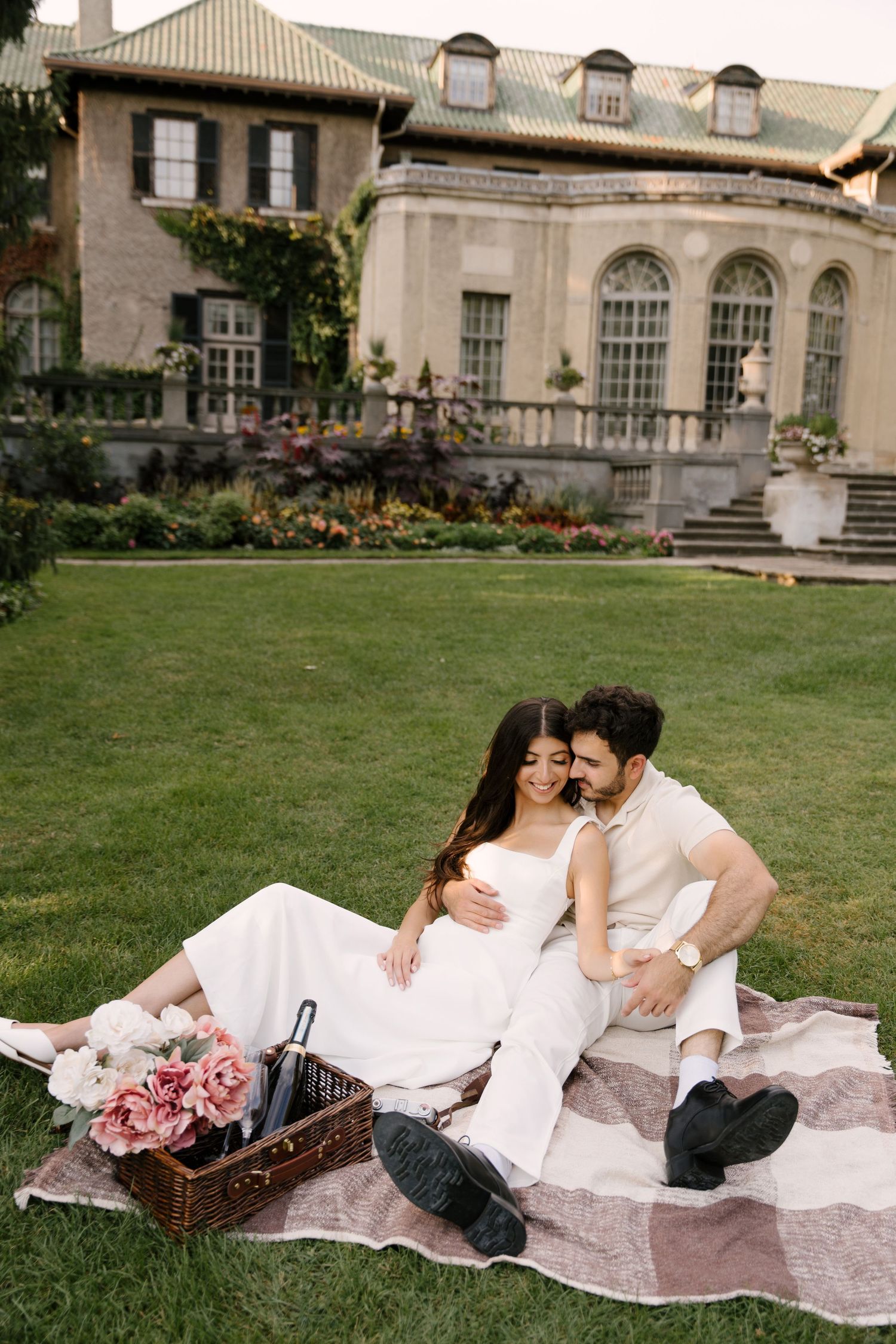 Couple sharing a romantic picnic on a blanket with flowers and wine basket on a lush estate lawn.