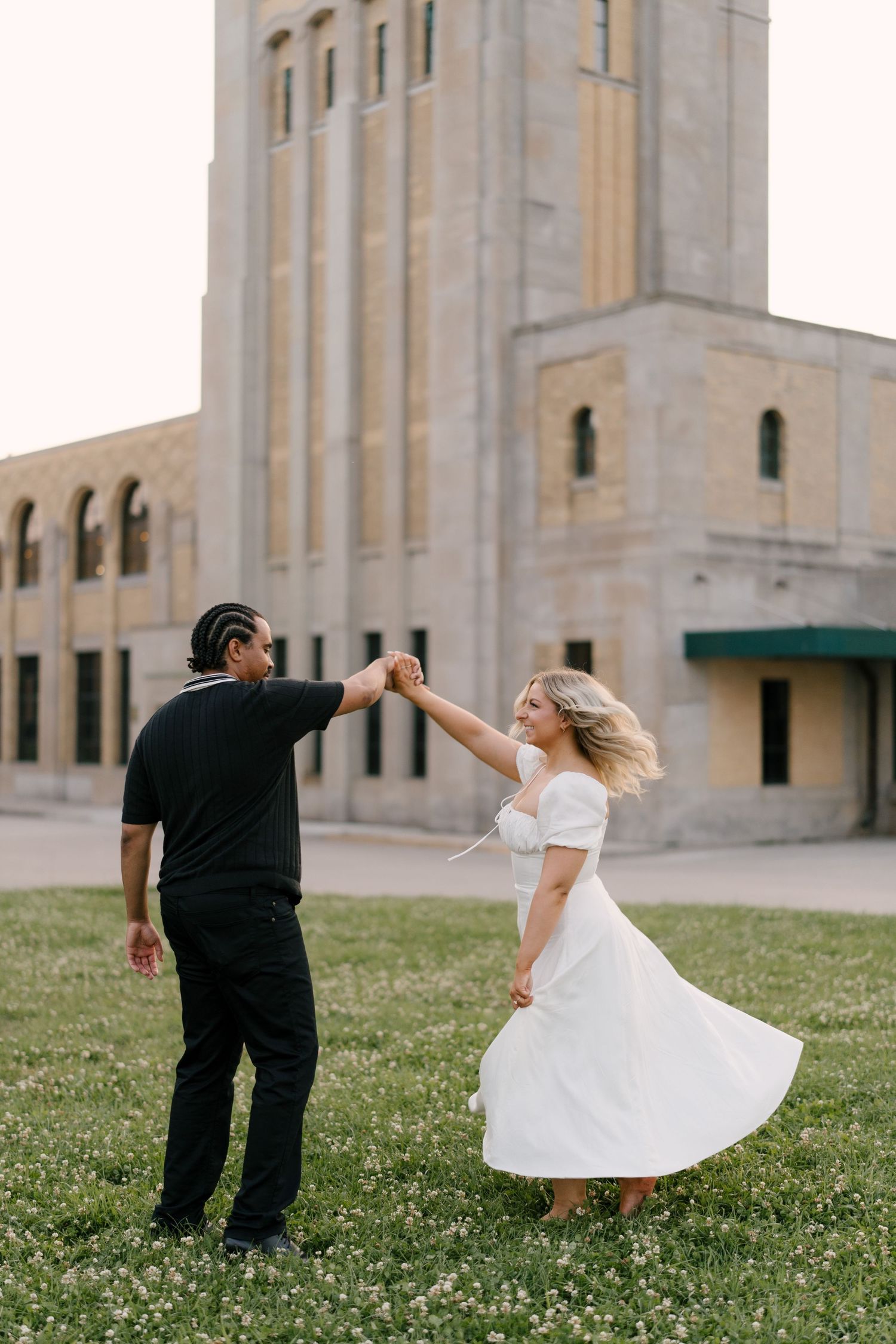 A man spins a laughing woman in a white dress on a green lawn in front of a large stone building.