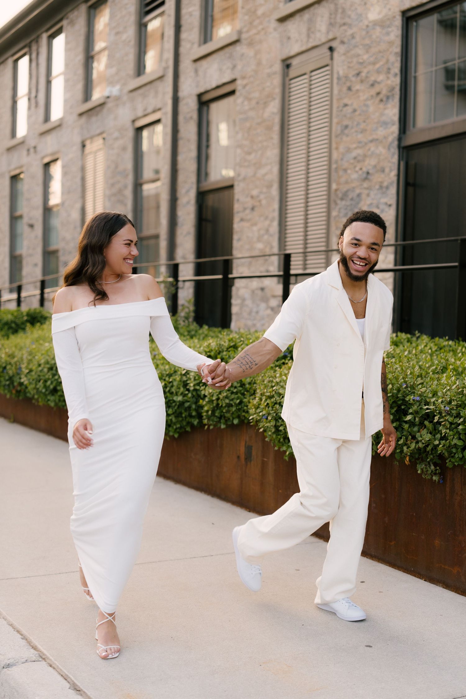 Joyful couple in white outfits hold hands and walk together outside a brick building.