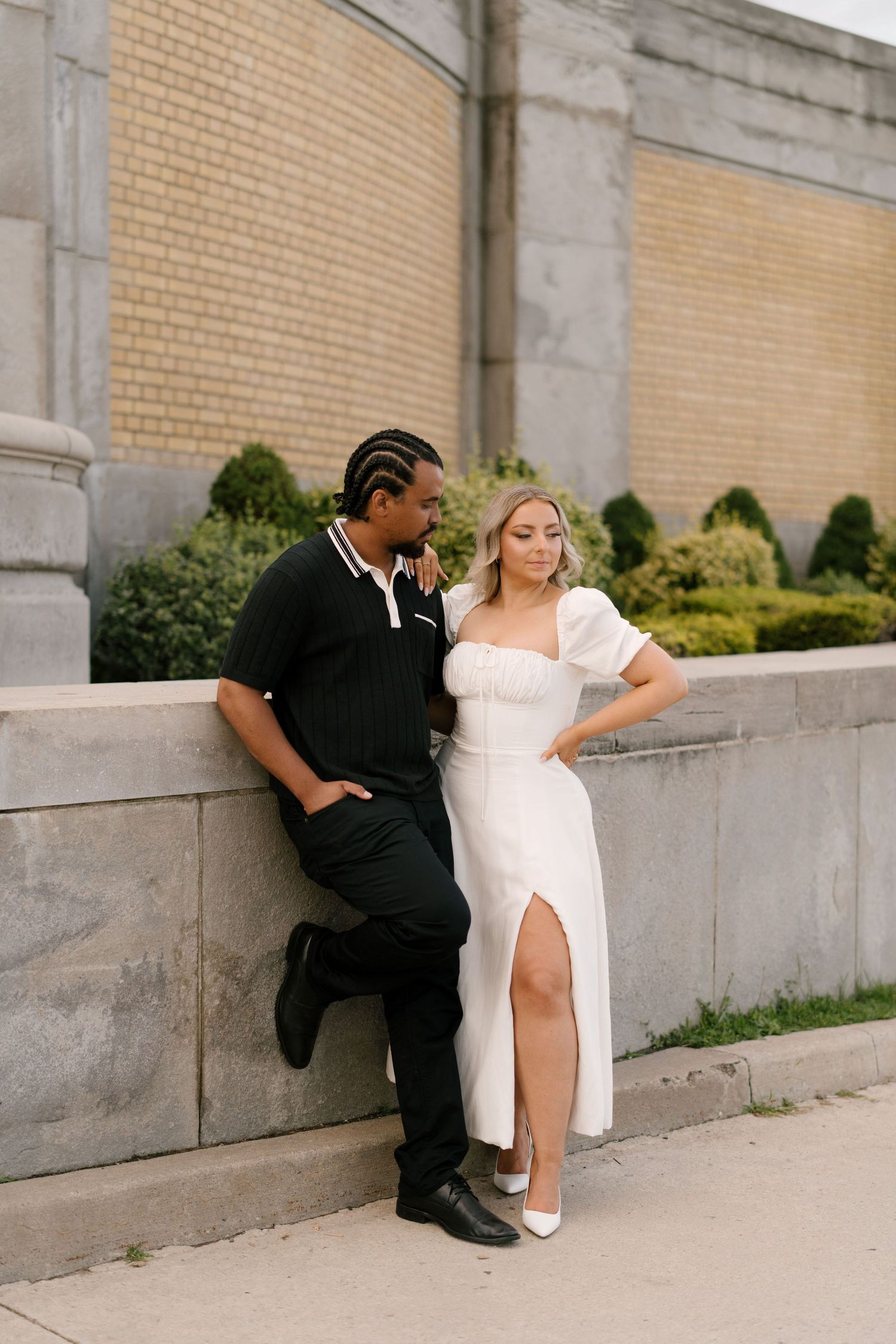 Stylish couple leaning against stone wall outdoors, woman in white slit dress and man in black outfit.