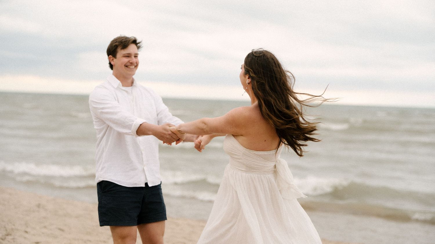 Joyful couple holding hands and spinning on a breezy beach, woman in flowing white dress.