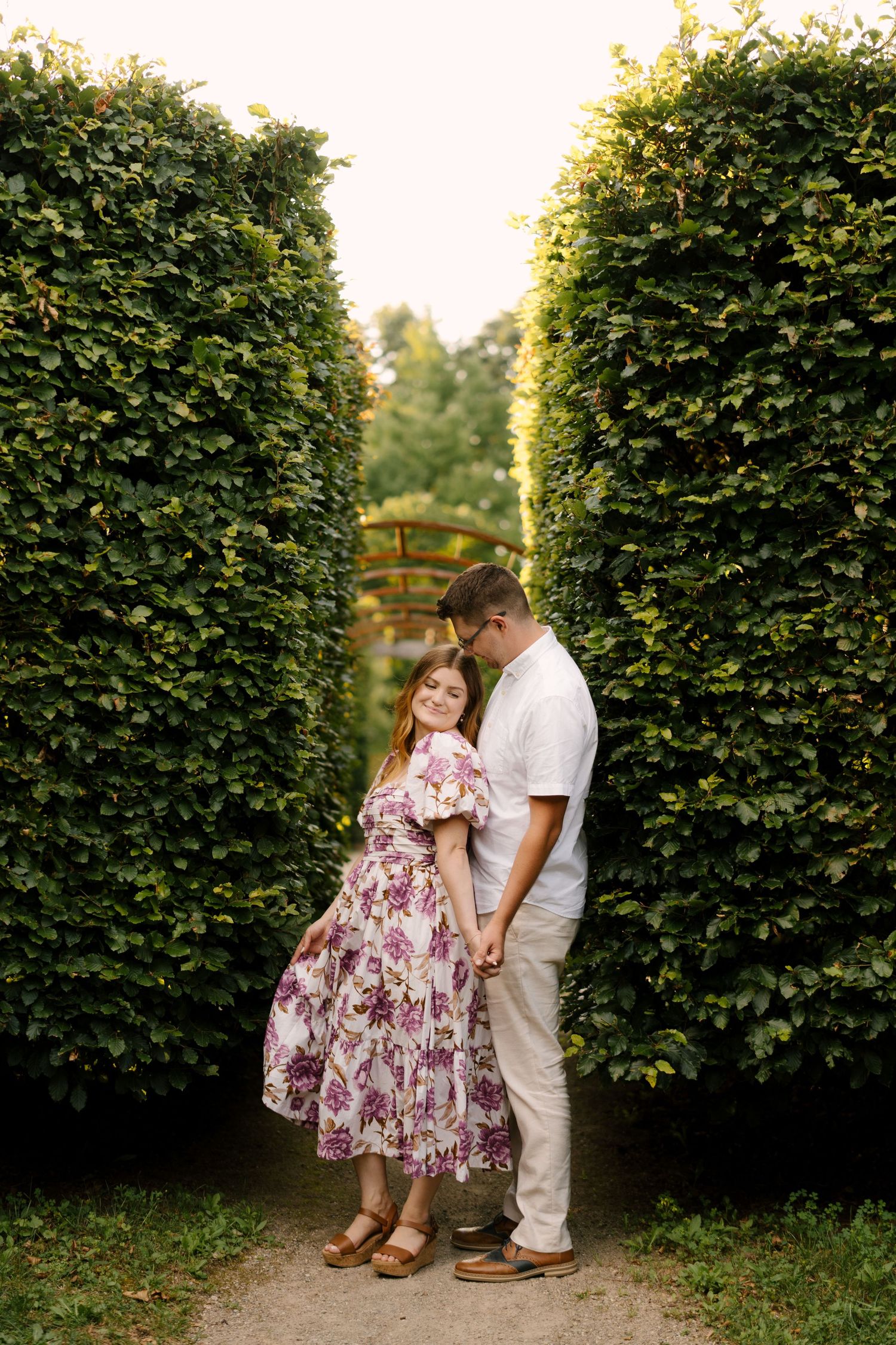 Romantic couple holding hands between tall green hedgerows in a lush garden pathway.