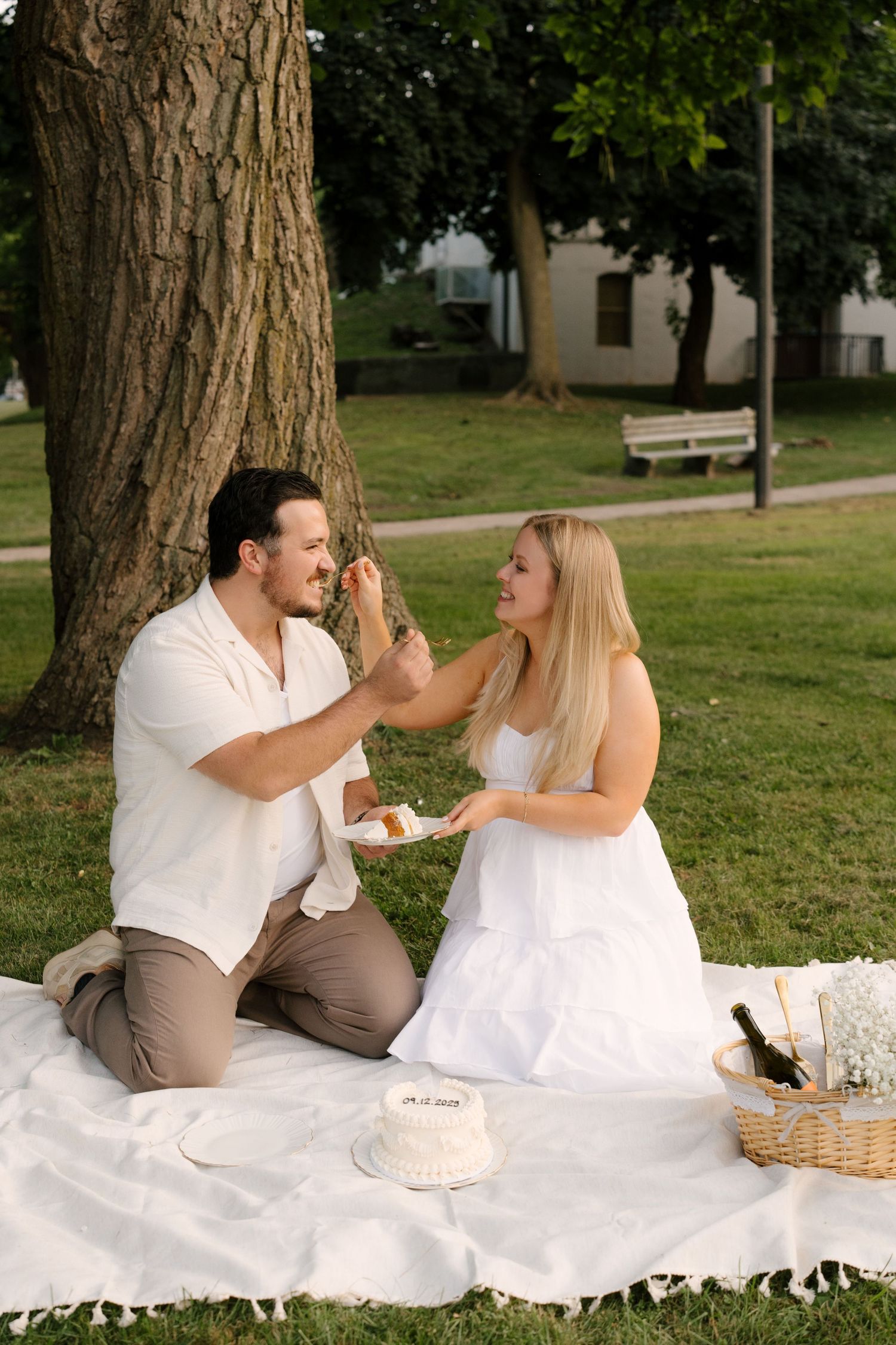 Woman feeding cake to a man on a white picnic blanket under a tree in a sunny park setting.