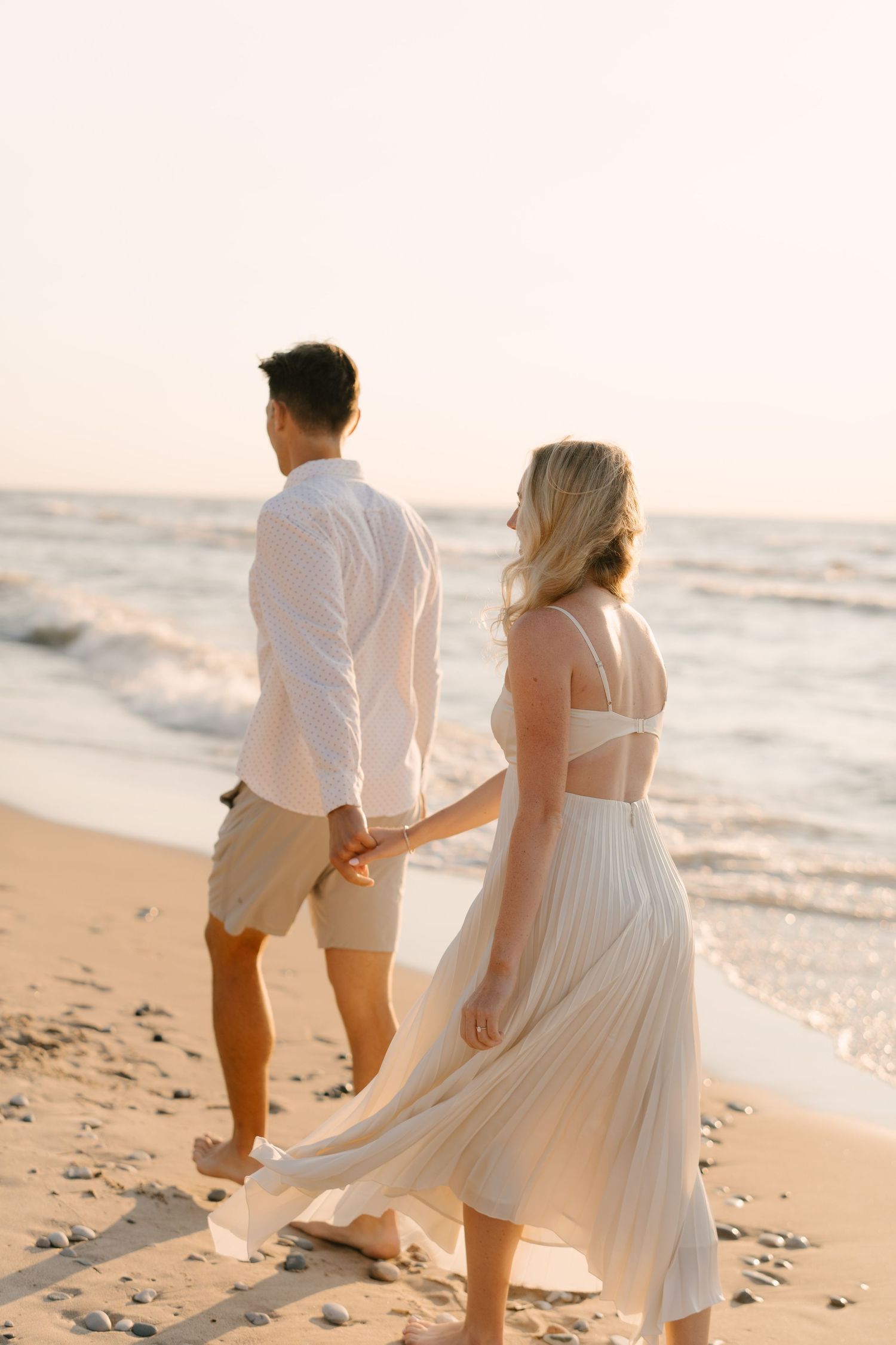 Couple holding hands walking barefoot on sandy beach at golden hour with ocean waves behind.