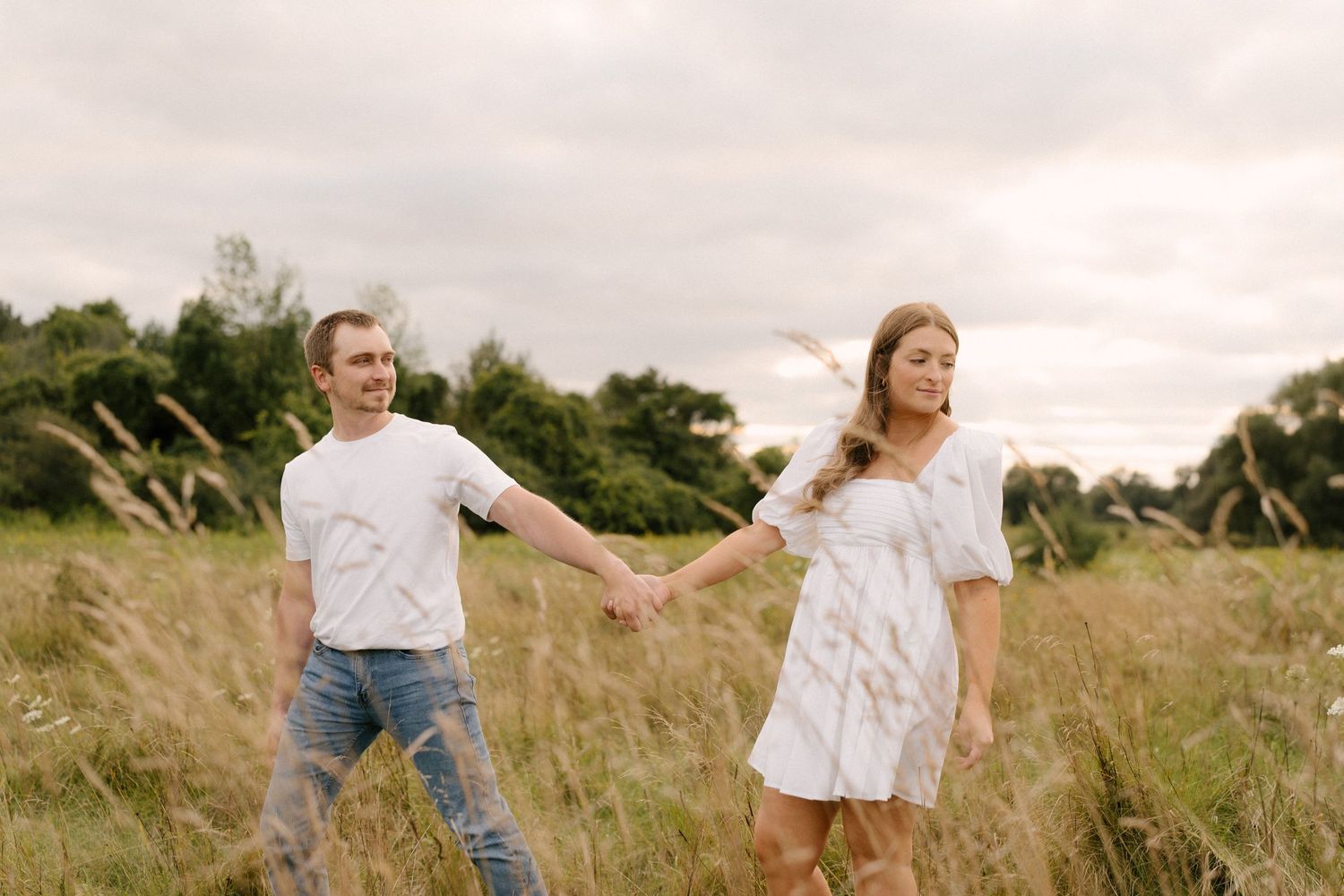 Happy couple holding hands while walking through tall grass in a scenic outdoor field setting.