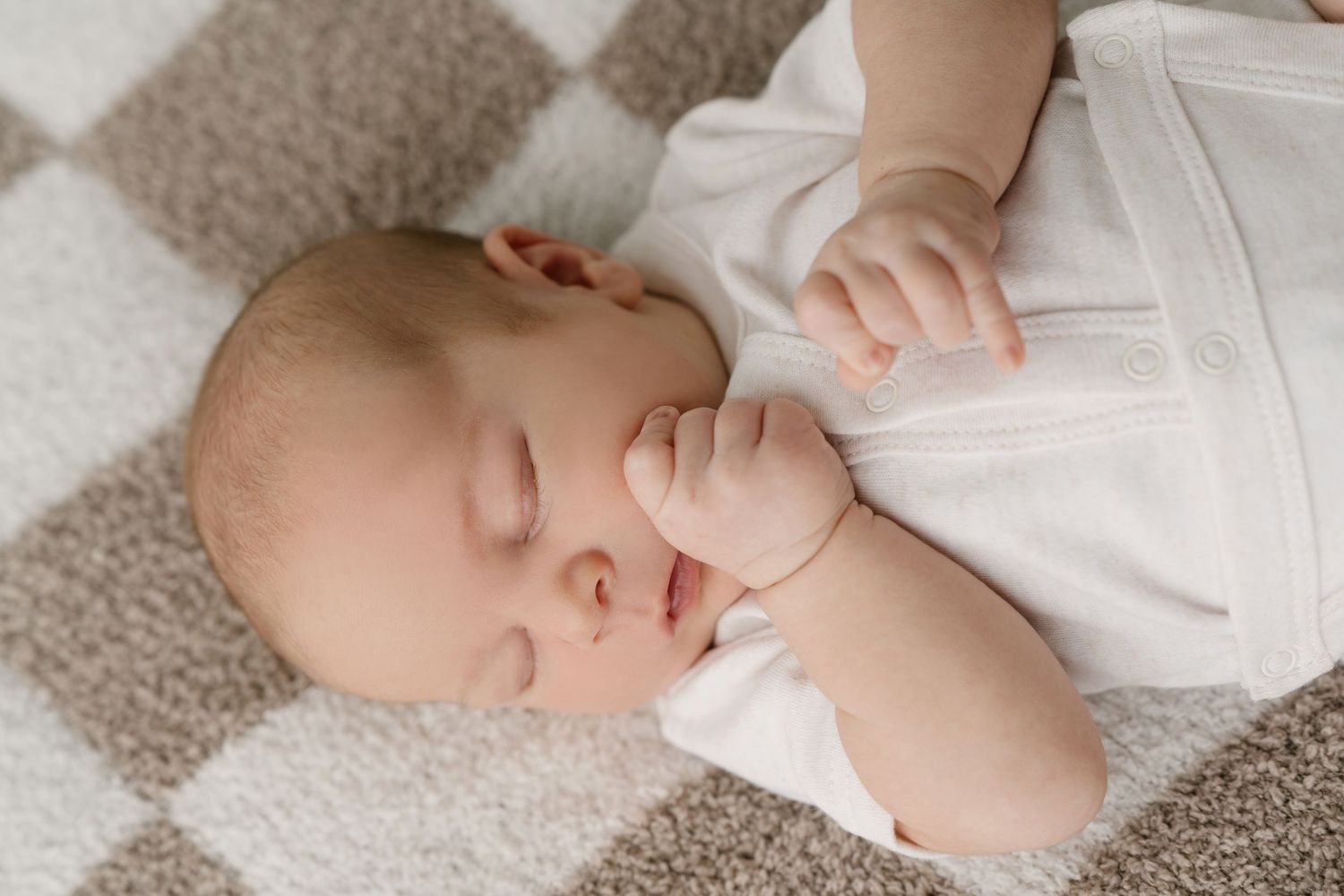 Sleeping newborn baby in white onesie lying on checkered blanket with hand near face.