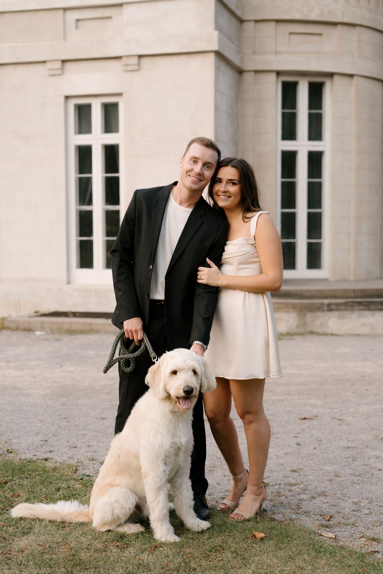 Smiling couple posing with large white golden doodle dog in front of elegant stone building.