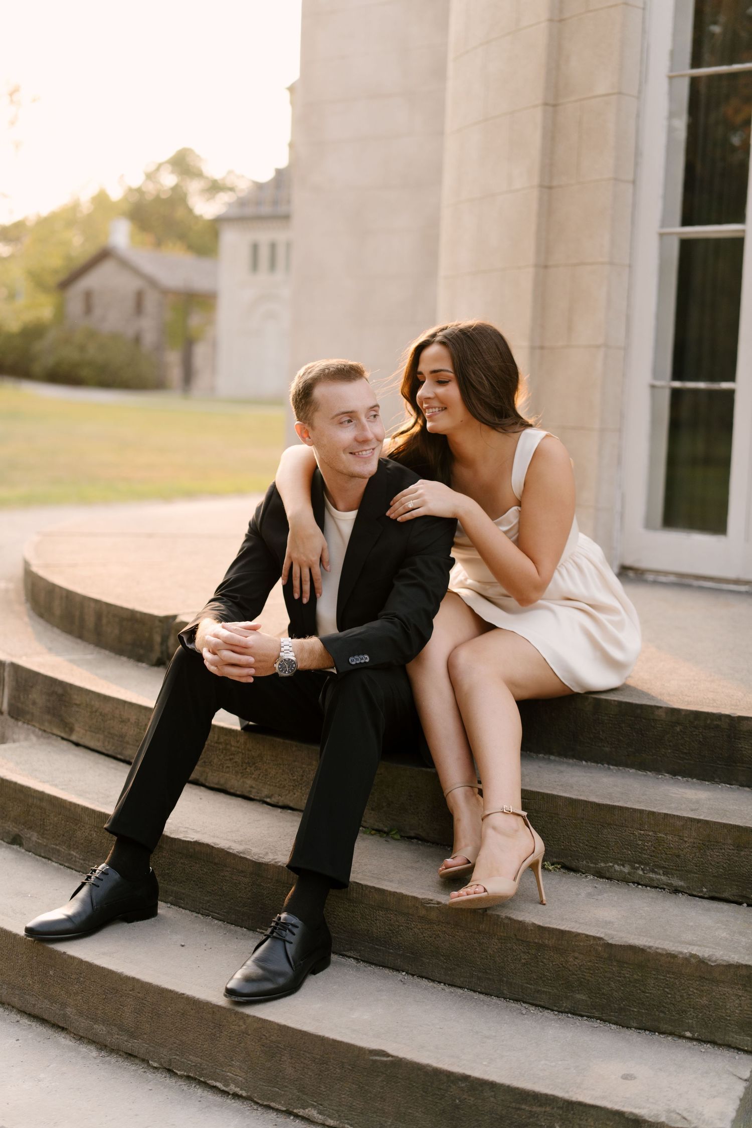 Couple in elegant attire sitting on stone steps, woman leaning into man affectionately.