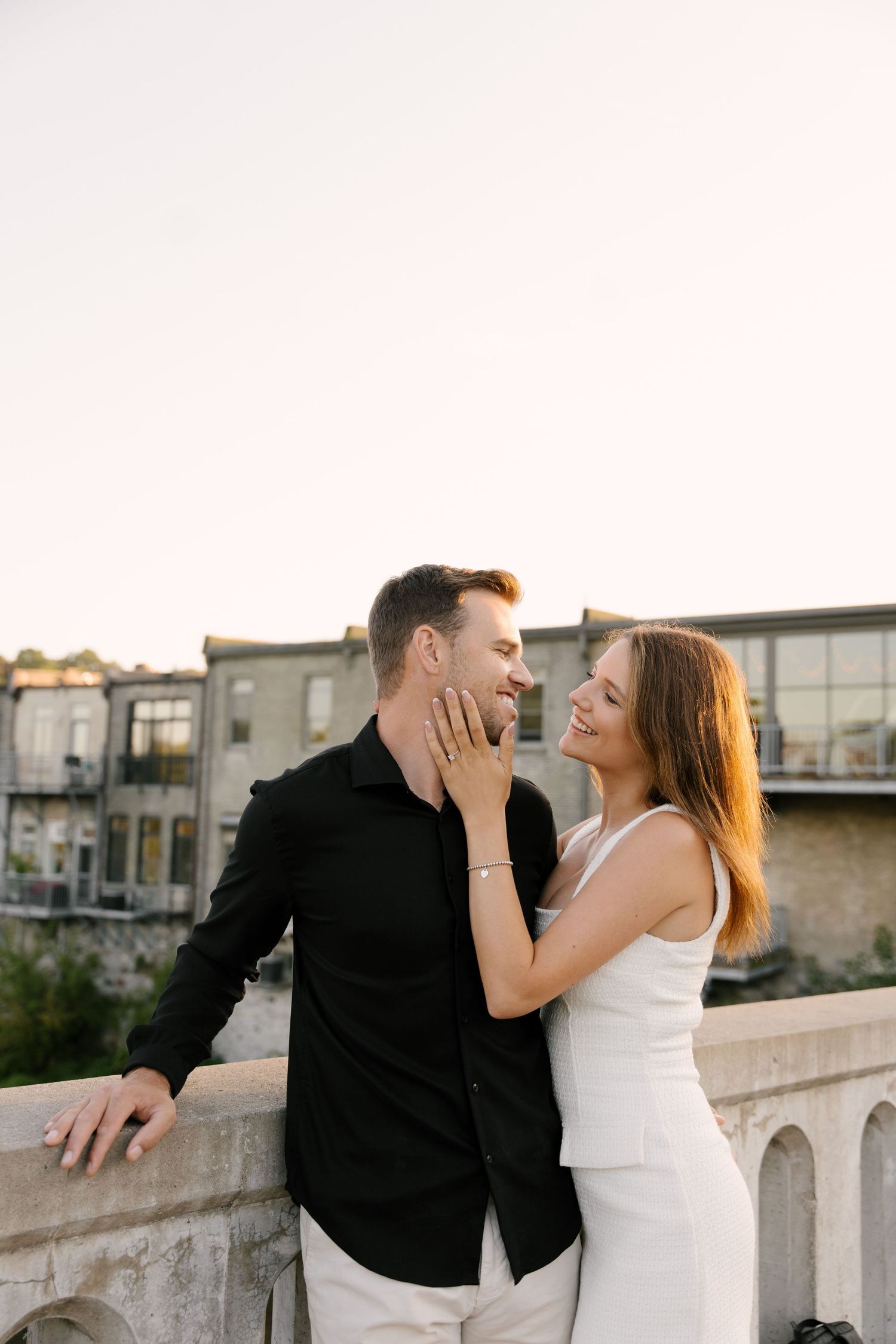 Happy couple laughing together on stone bridge with urban buildings behind.