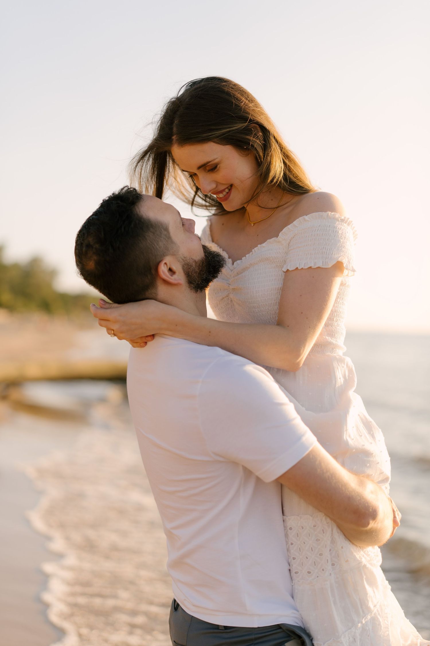 Couple sharing a tender moment at the beach, man lifting smiling woman in white off-shoulder dress at golden hour.