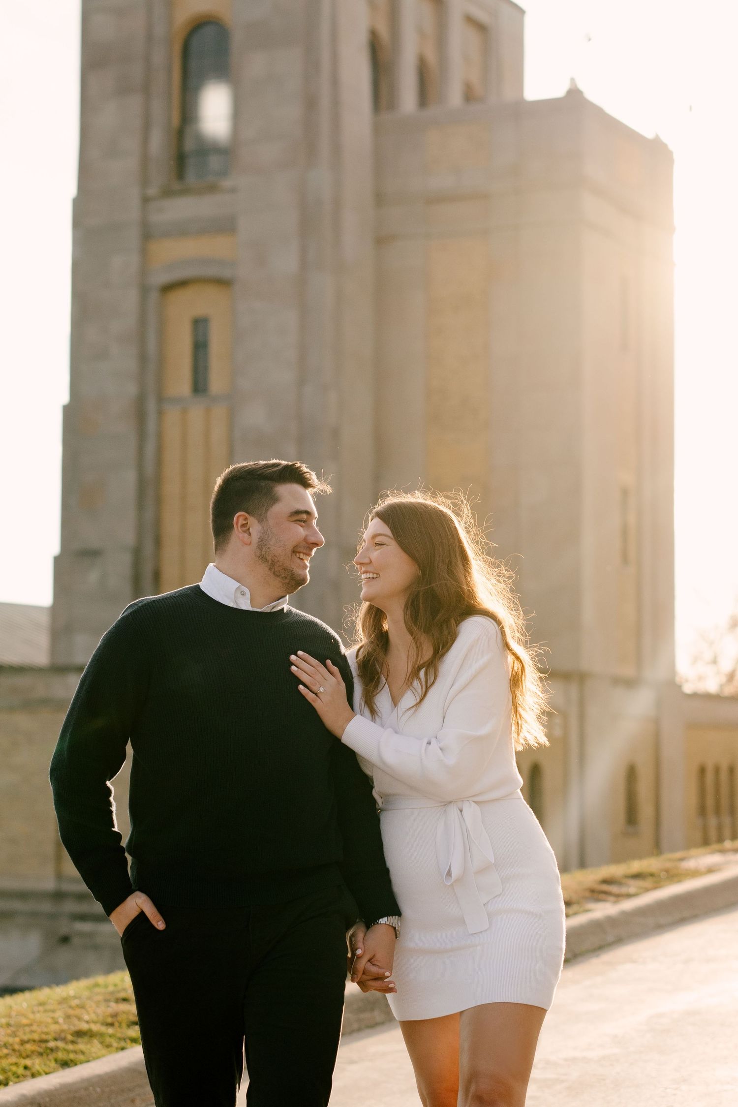 Happy couple holding hands and smiling at each other with sunlit tower behind them.