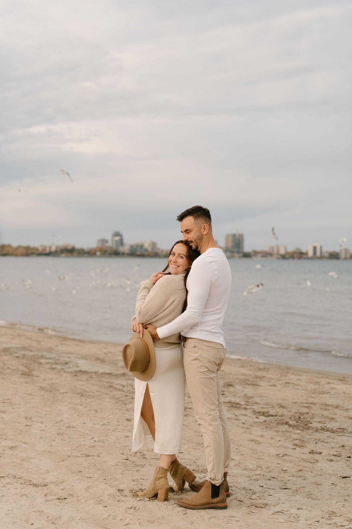 Couple embracing on sandy beach with city skyline and seagulls in background.