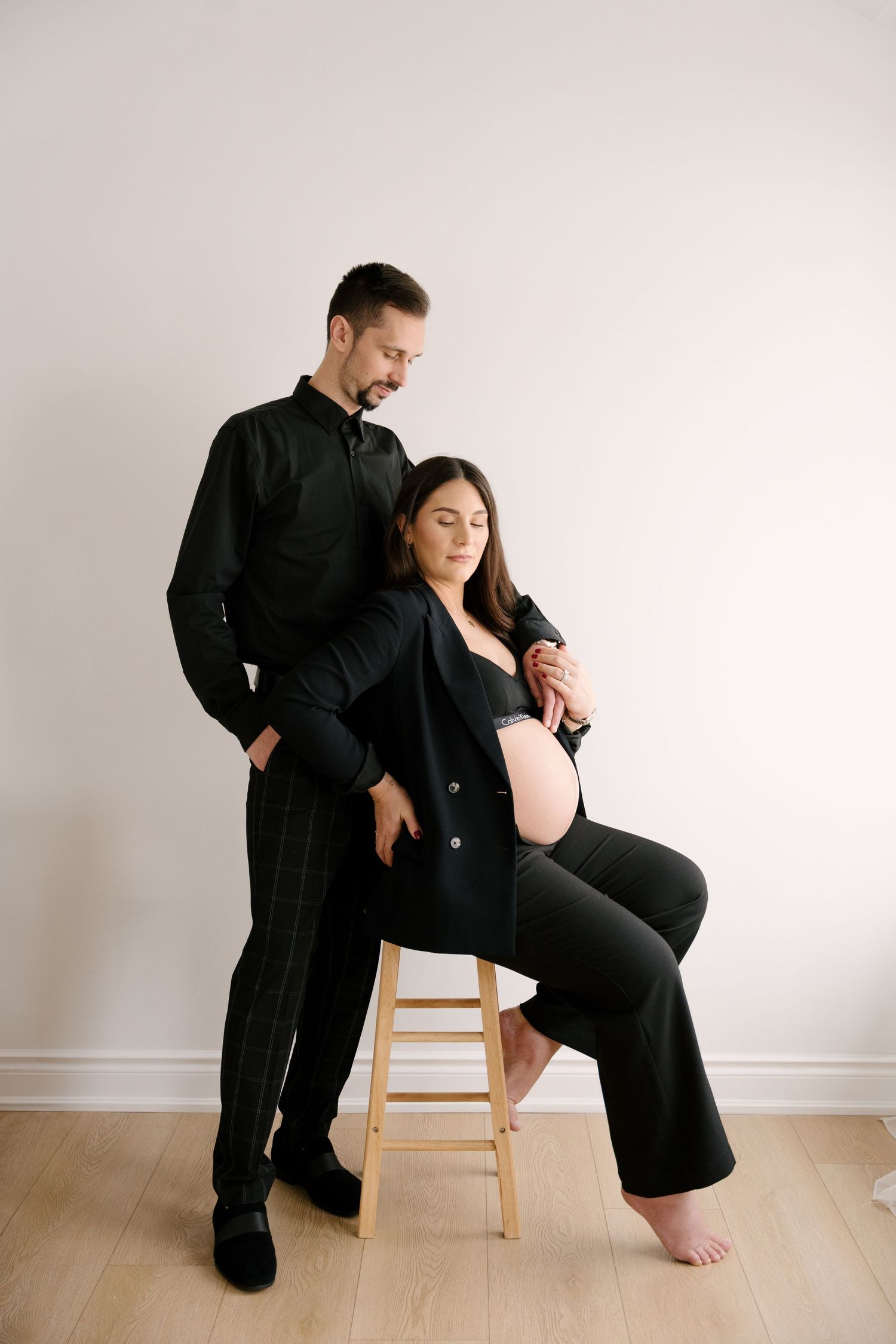 Pregnant woman in black blazer seated on stool with partner standing behind her in a bright studio.