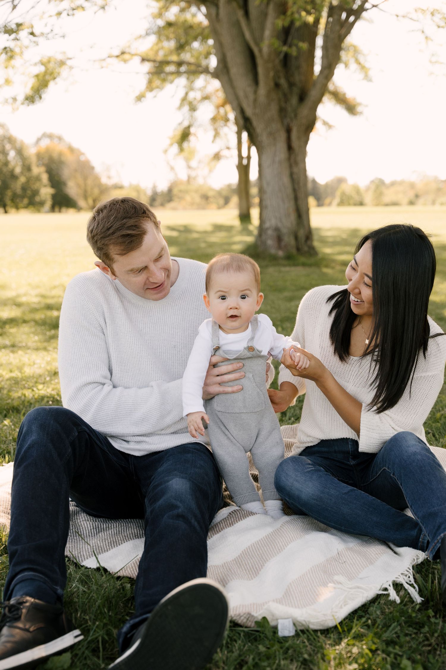 Family with baby on blanket in sunny park, parents smiling at infant in gray overalls.