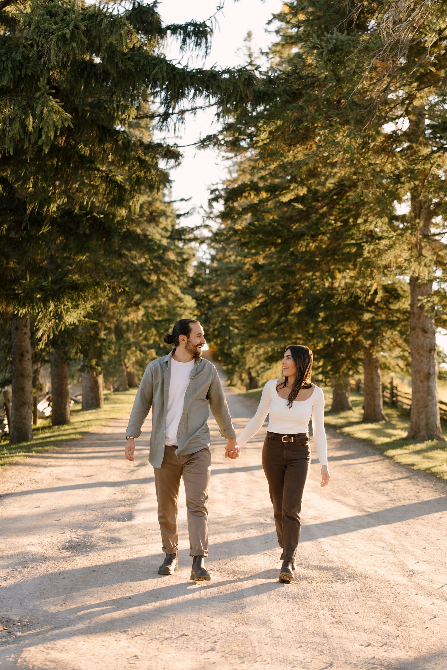 Couple holding hands and walking along a sunlit path lined with tall evergreen trees.