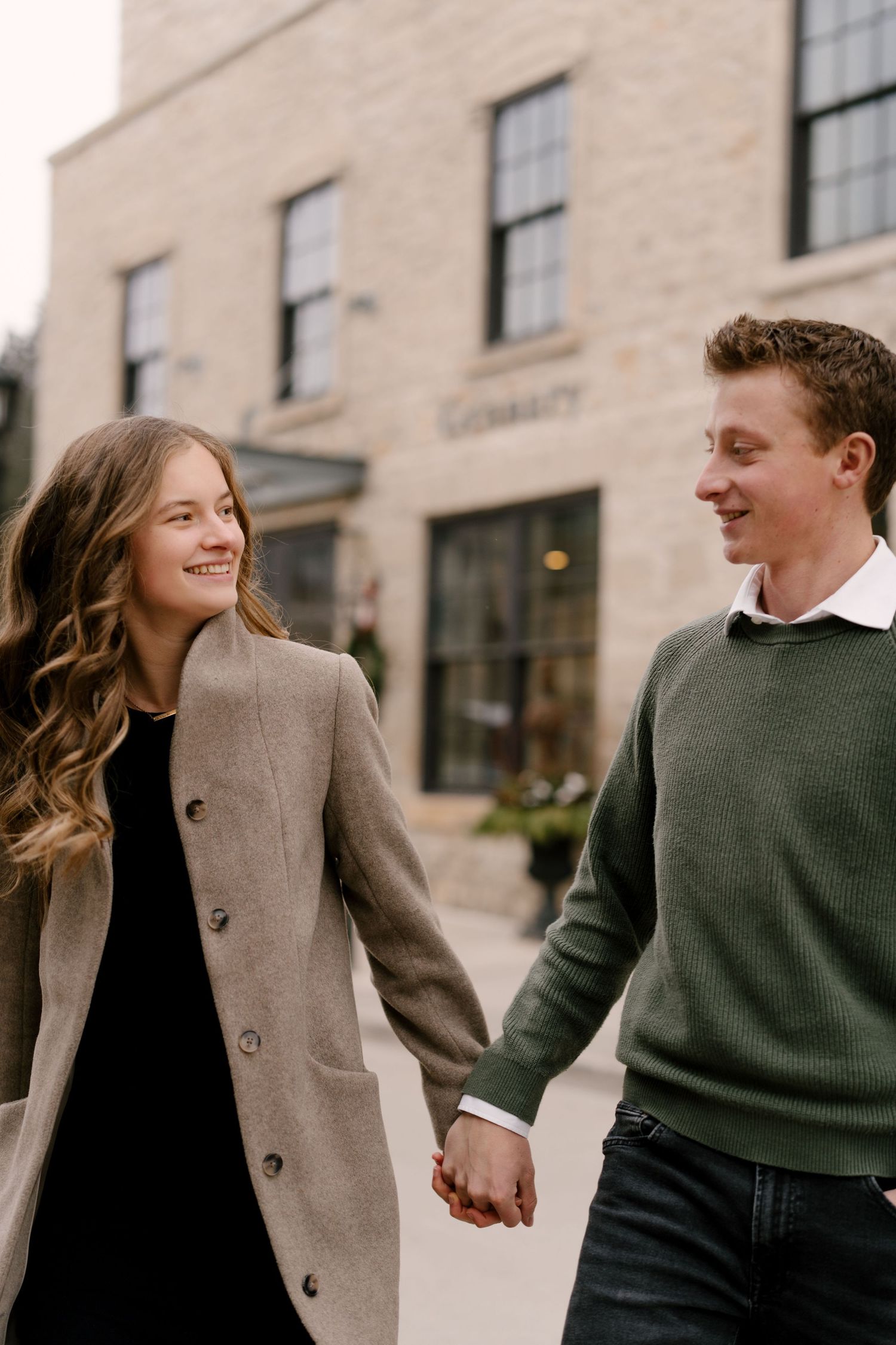 A smiling couple holds hands while walking past a stone building on a charming street.