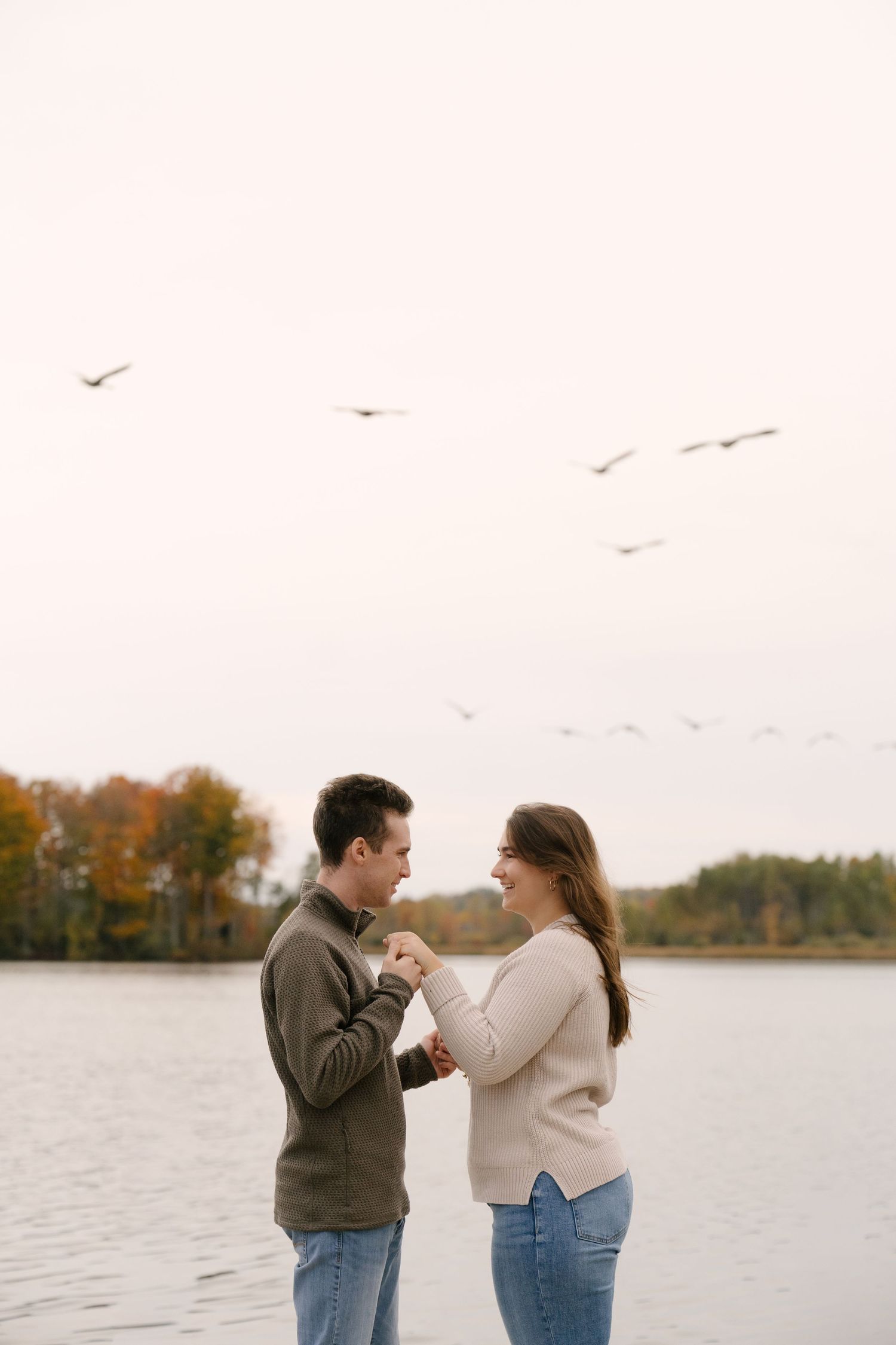 Couple holding hands by calm lake, birds flying overhead, autumn trees reflecting in water.