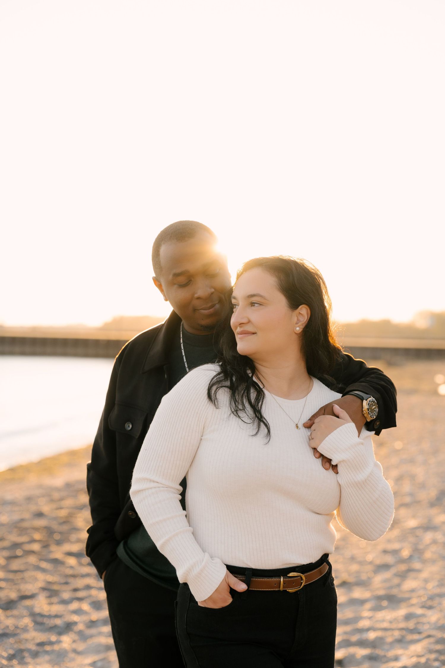 Interracial couple standing together on a sandy beach at golden hour, smiling warmly.