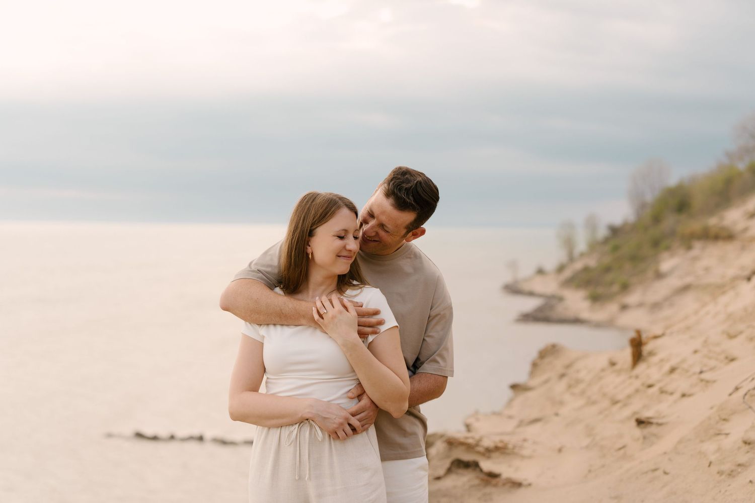 Couple embracing on sandy beach cliff with calm ocean backdrop in soft natural light.