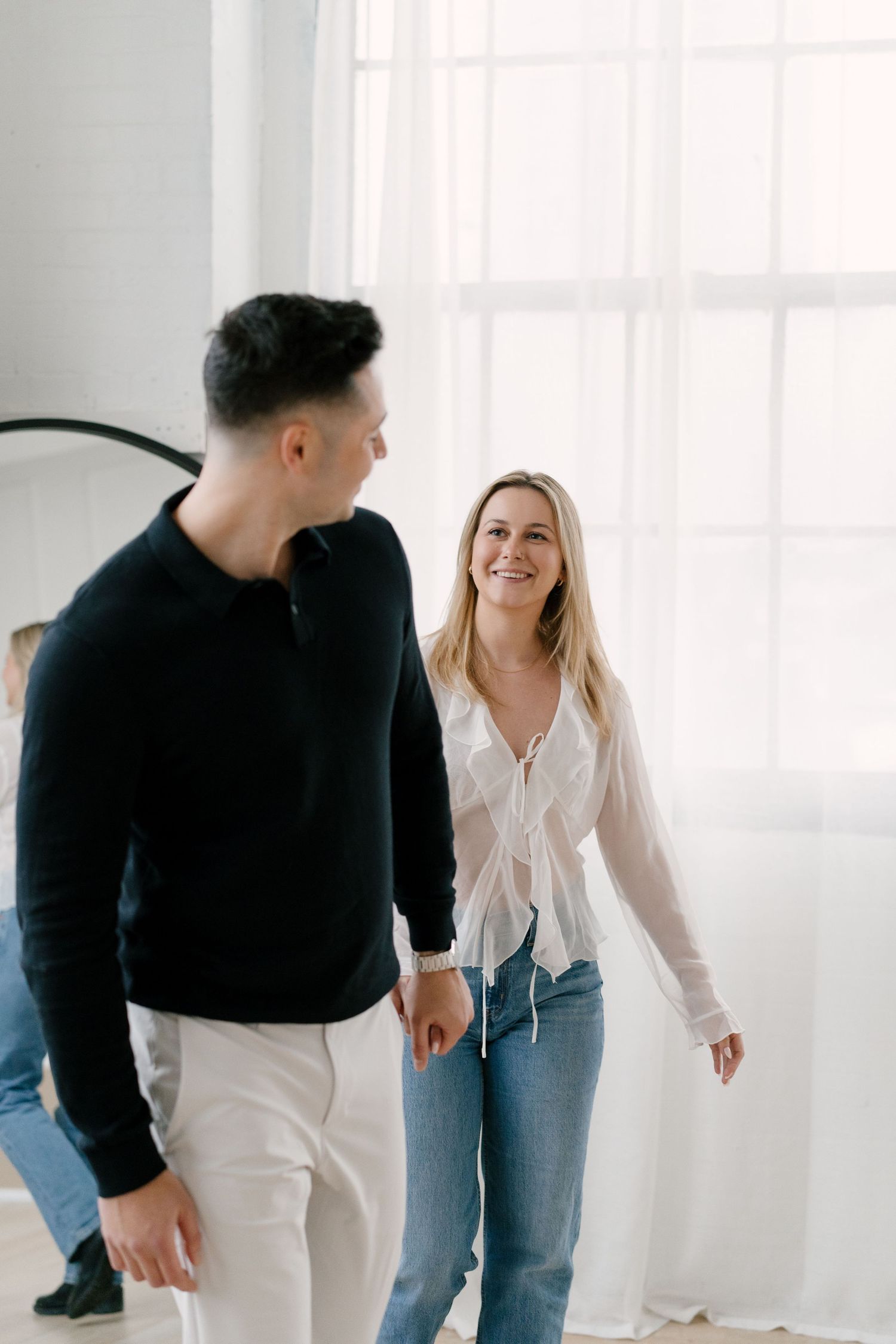 Smiling blonde woman walking with a dark-haired man in a bright white sunlit room.