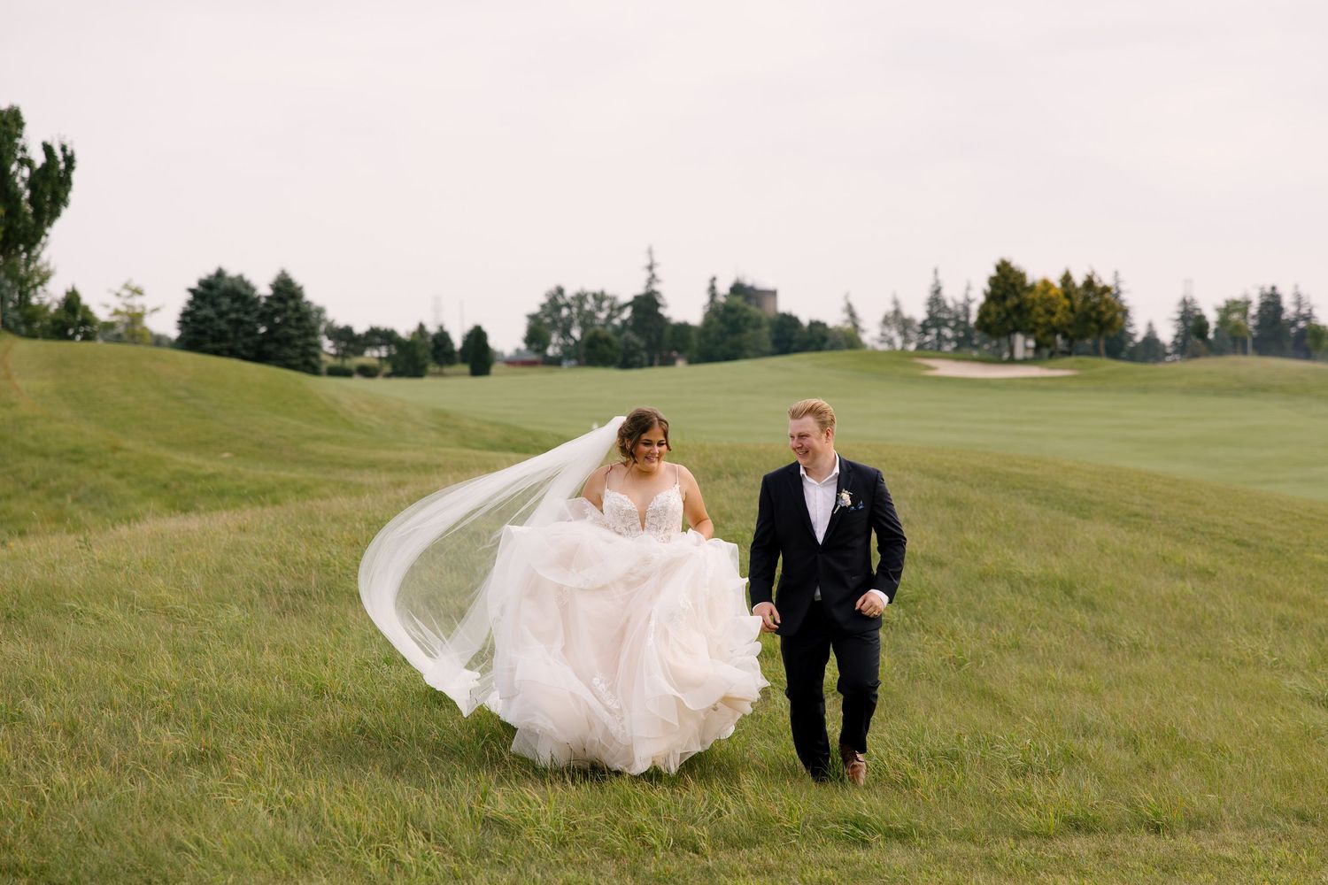 Bride and groom walk hand in hand on a lush green golf course, her veil flowing dramatically in the wind at Whistle Bear venue.