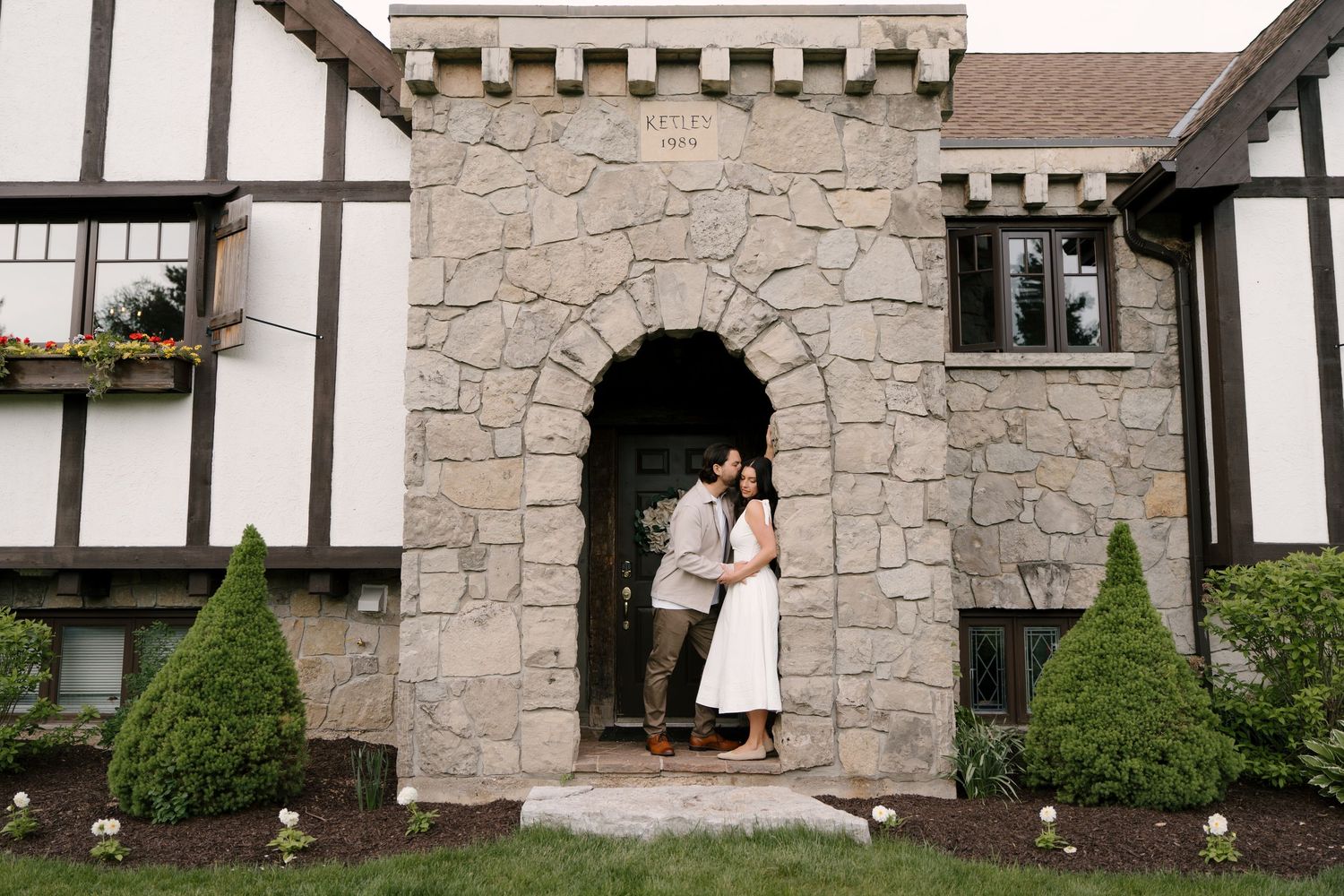 Couple sharing a romantic moment under a stone arch doorway of a Tudor-style building surrounded by manicured hedges.