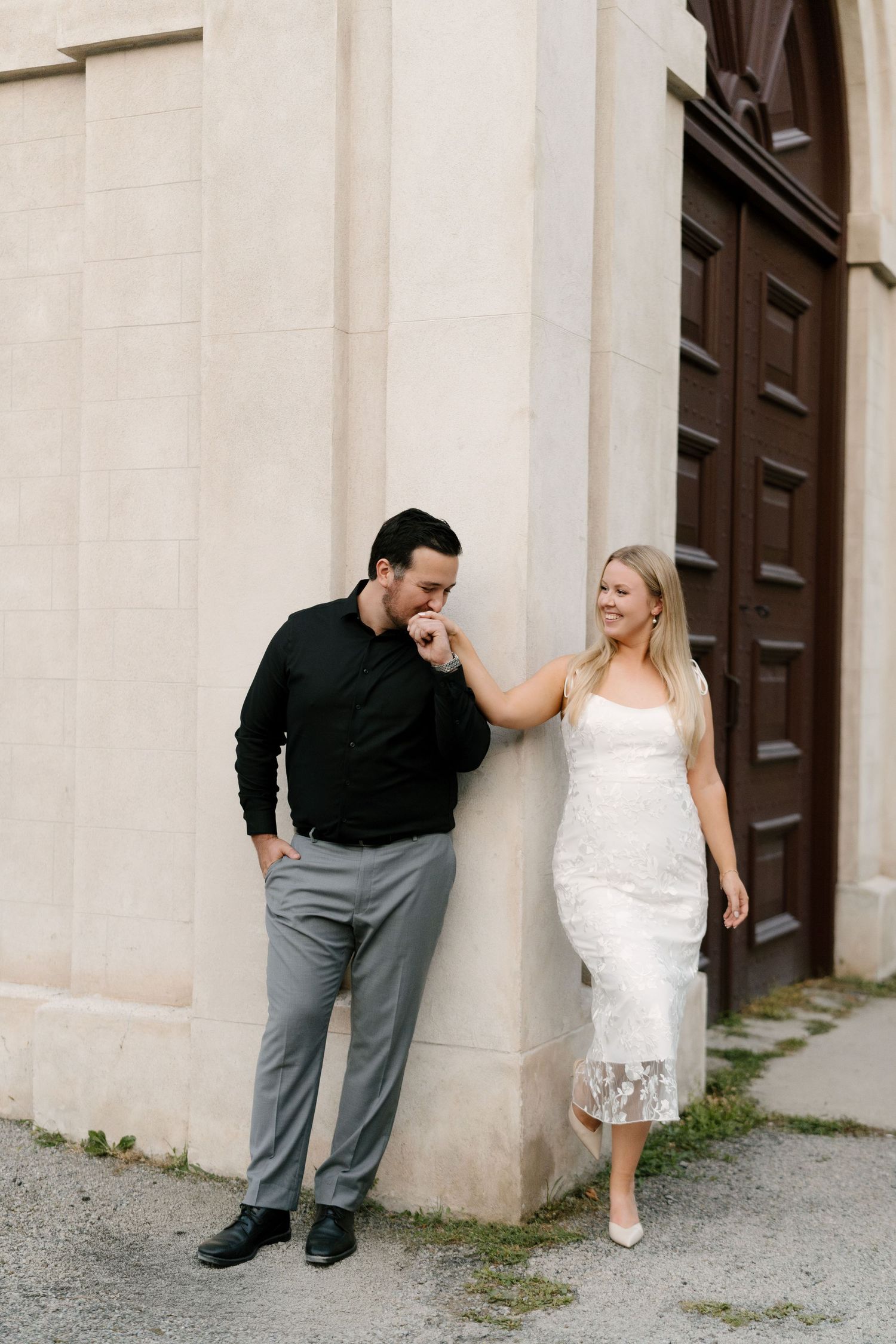 Man kissing womans hand while she smiles in a white lace dress against a stone building.