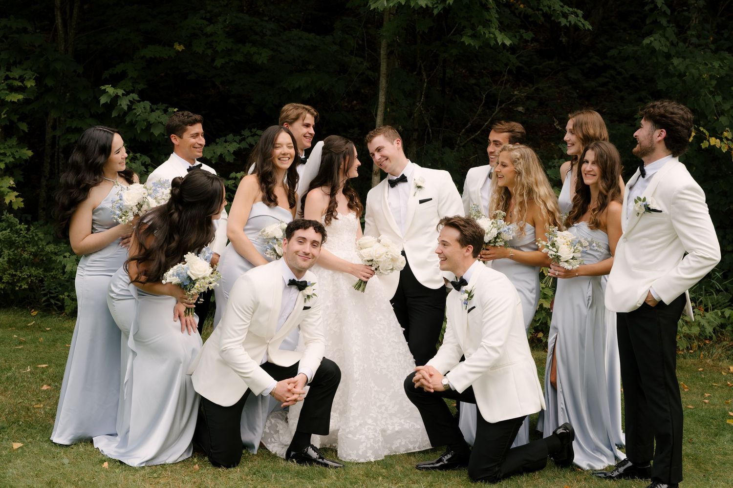 Wedding party in white and light blue attire posing outdoors on green lawn with bride and groom centered.