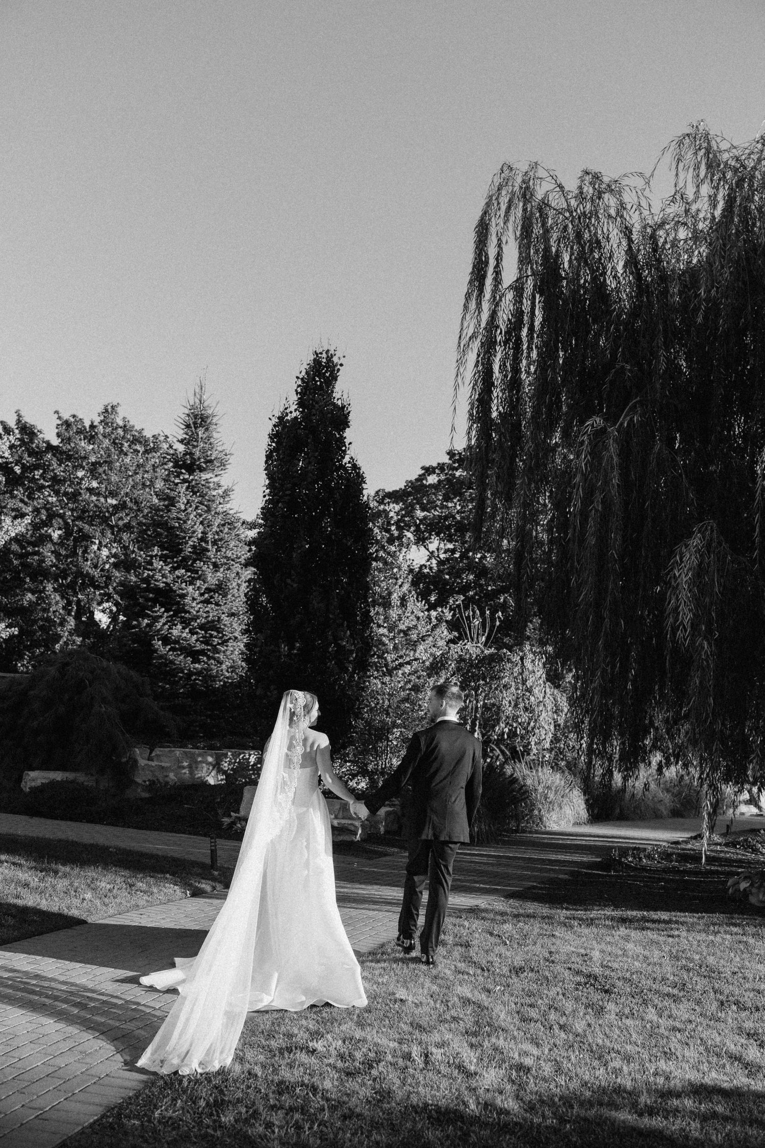 Bride and groom strolling through a scenic park with tall trees in black and white.