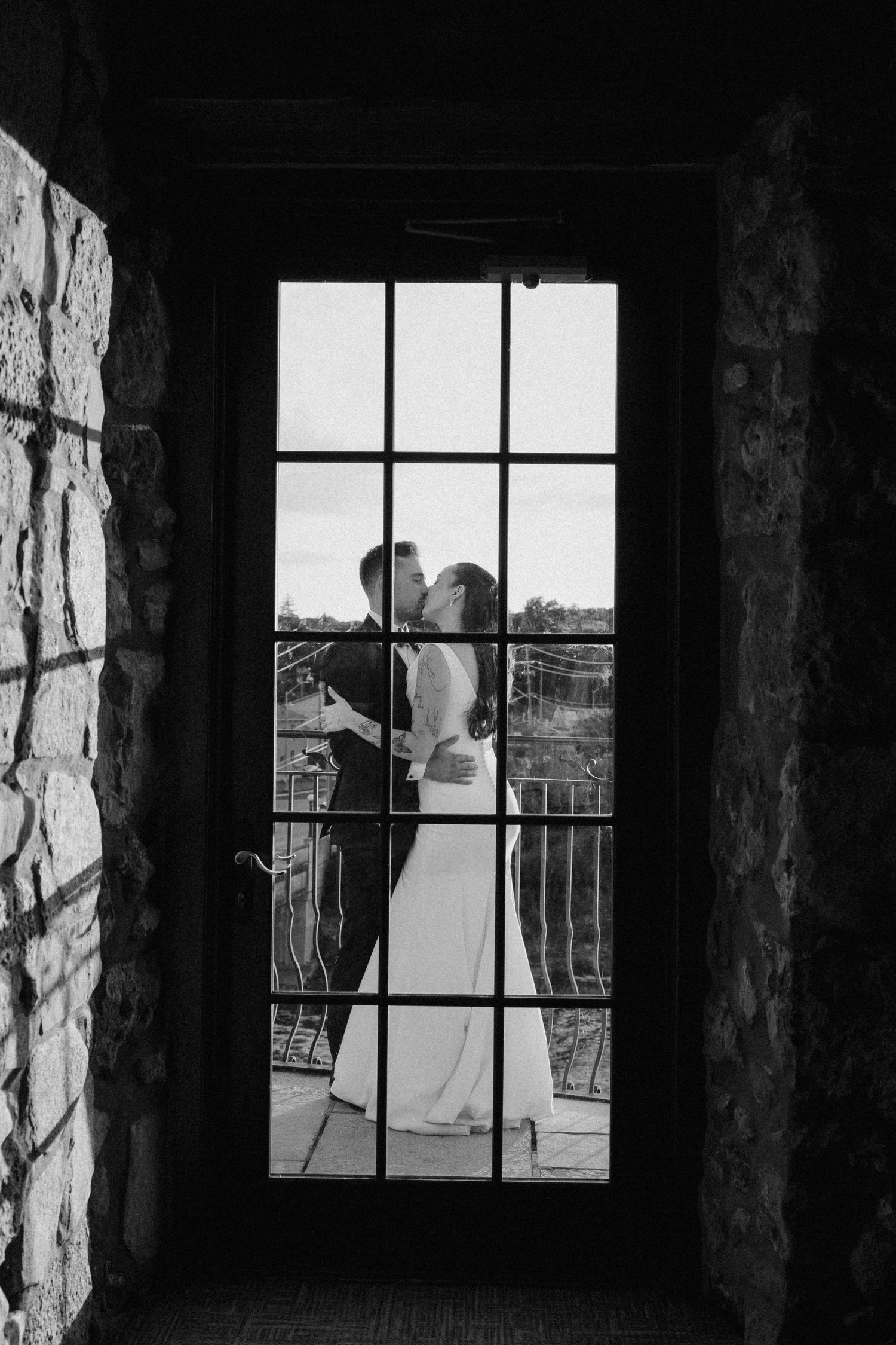 Bride and groom share romantic kiss framed by glass door with stone wall in black and white at Cambridge Mill.