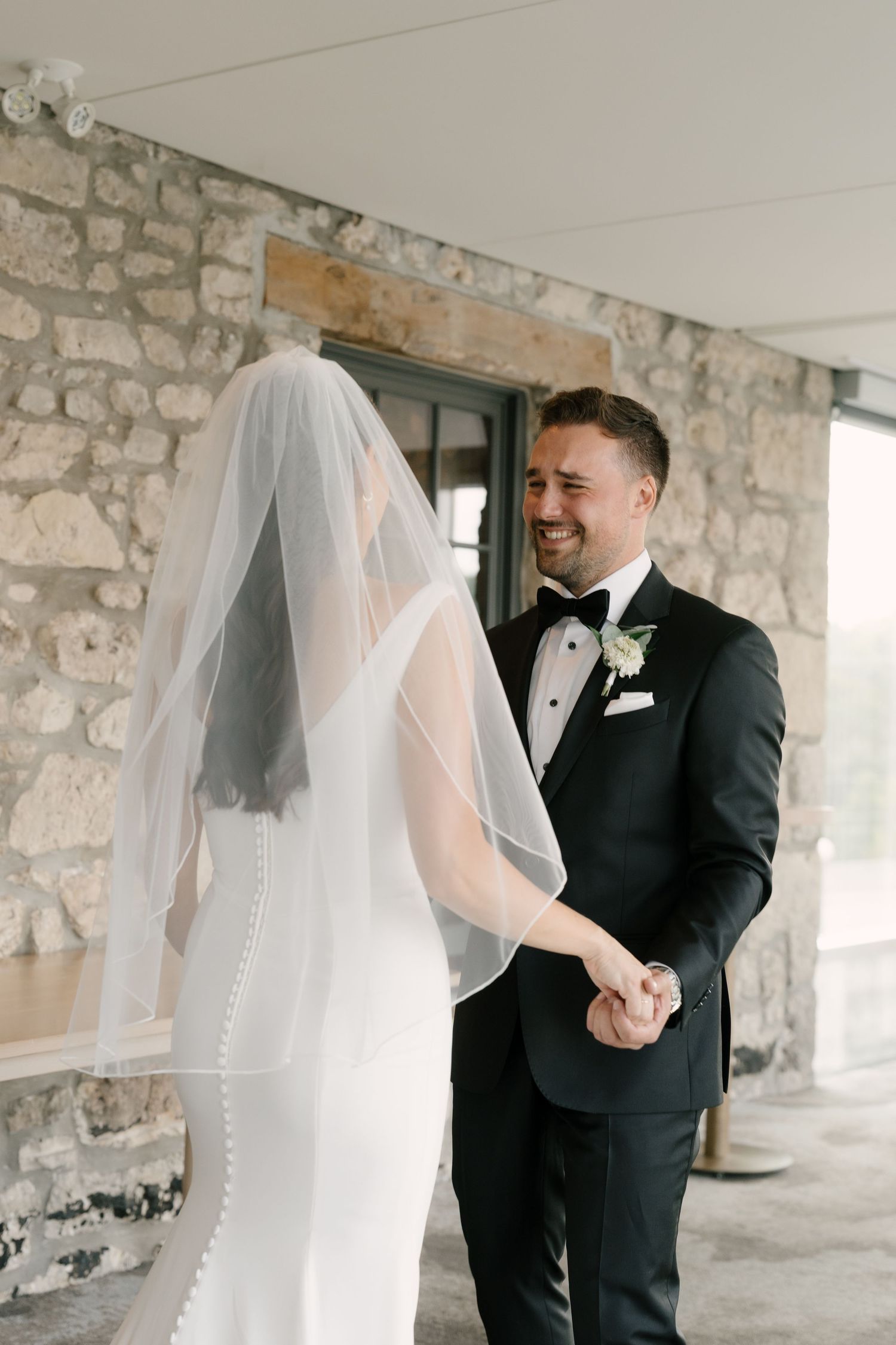 Groom in black tuxedo smiling at bride in white wedding dress and veil during first look at stone Cambridge Mill venue.