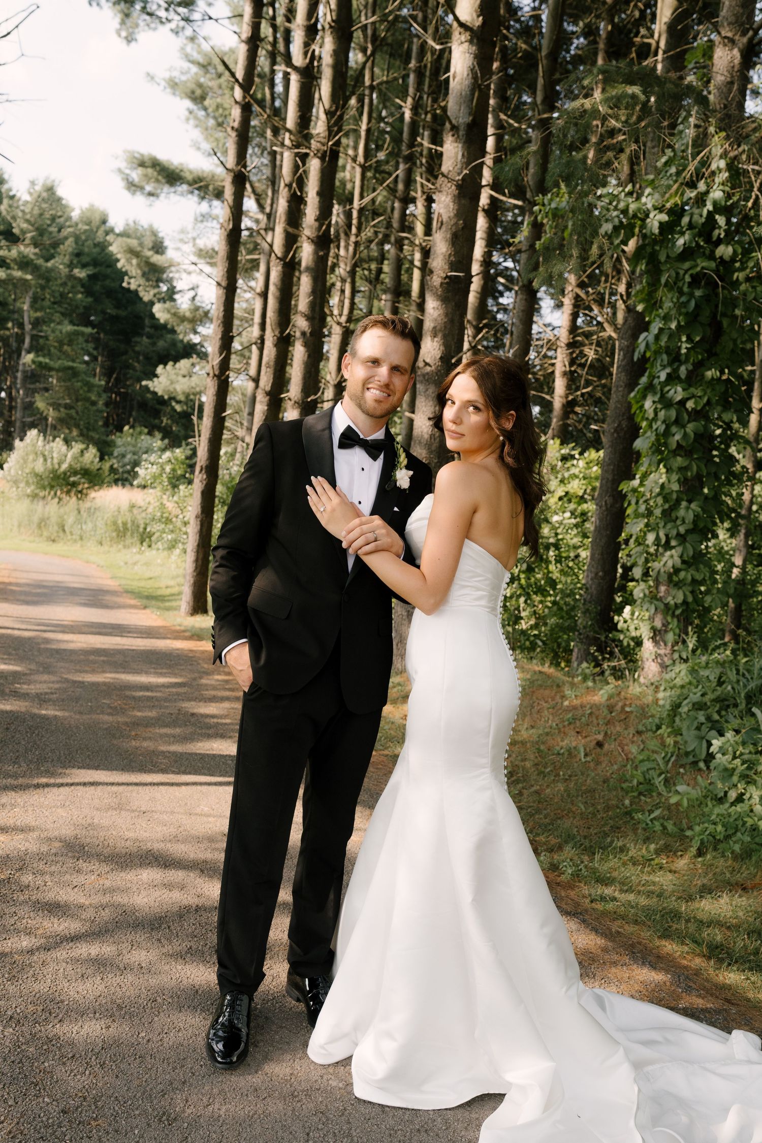 Bride and groom smiling together on a tree-lined path, she in a white mermaid gown, he in a black tuxedo.