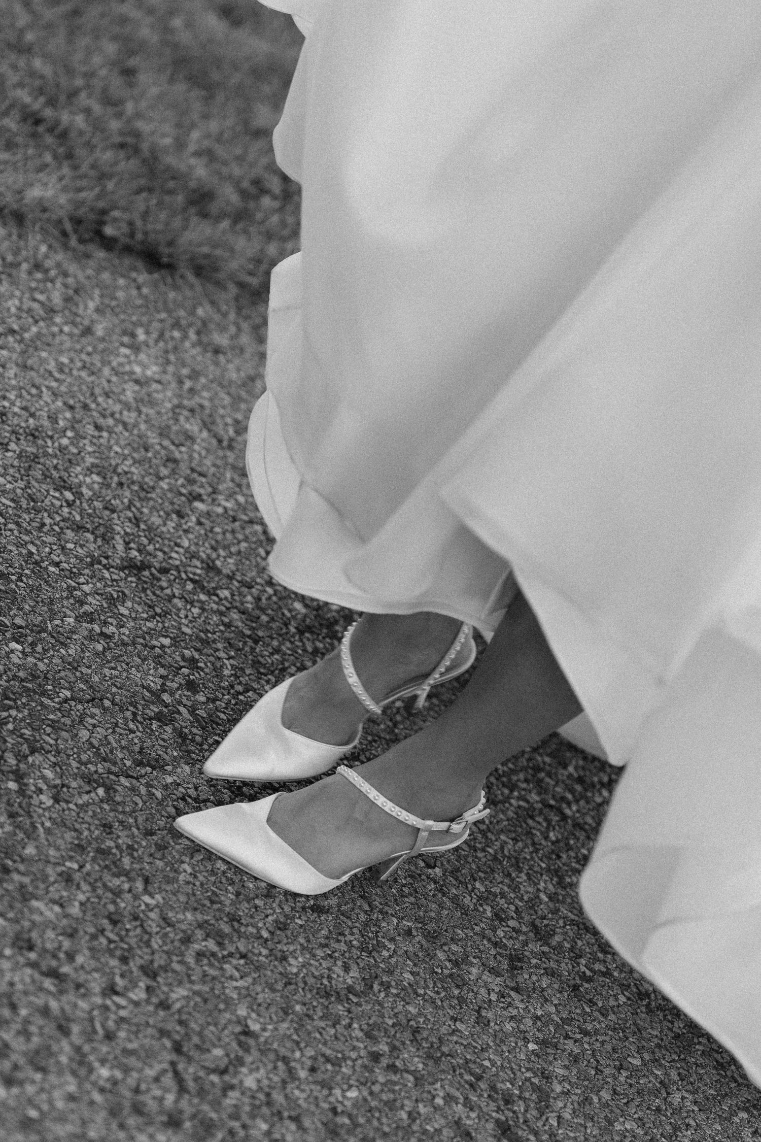 Close-up black and white photo of bride wearing pointed-toe satin heels with ankle straps beneath wedding gown.