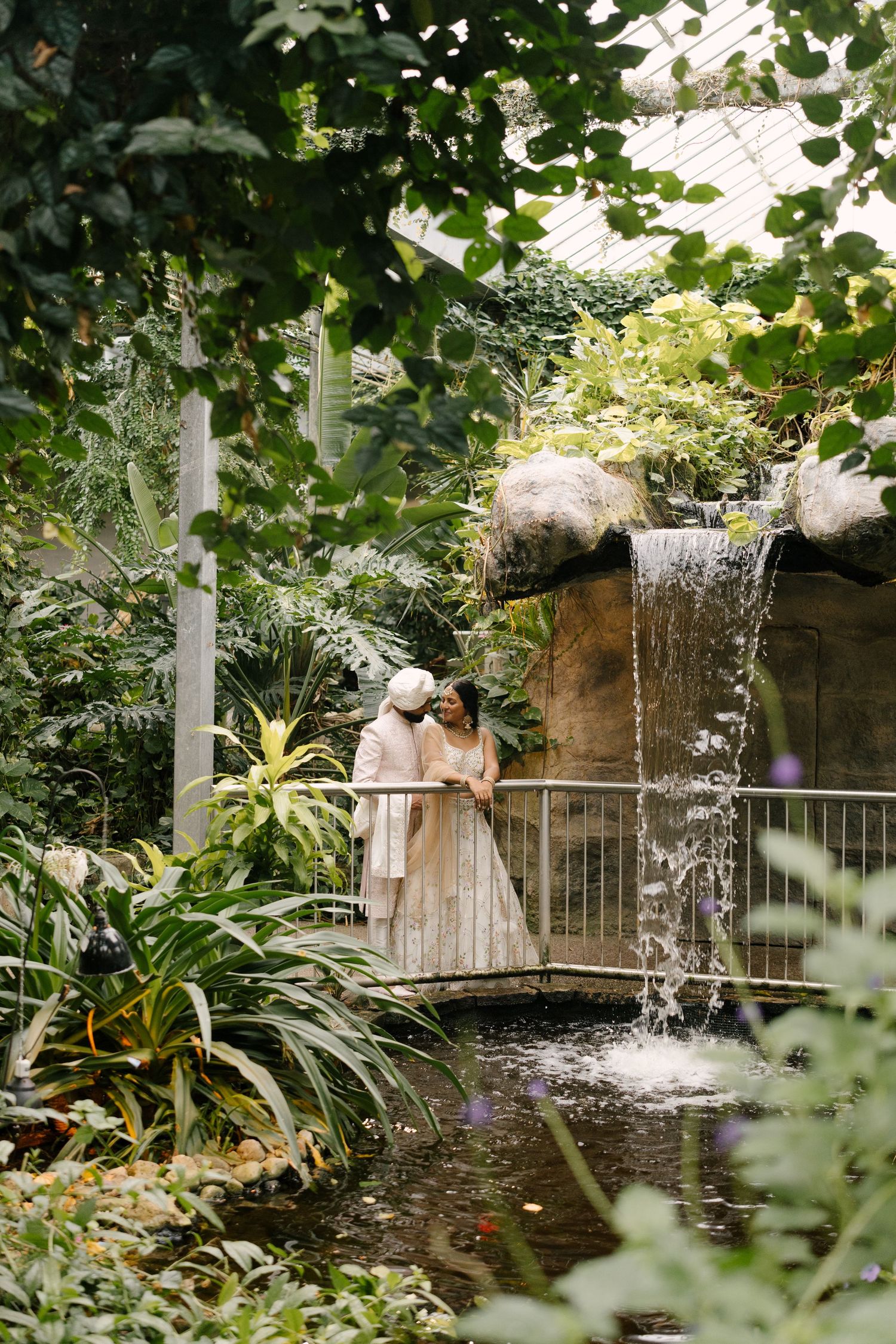 Indian wedding couple share intimate moment on bridge beside waterfall in tropical butterfly conservatory greenhouse.