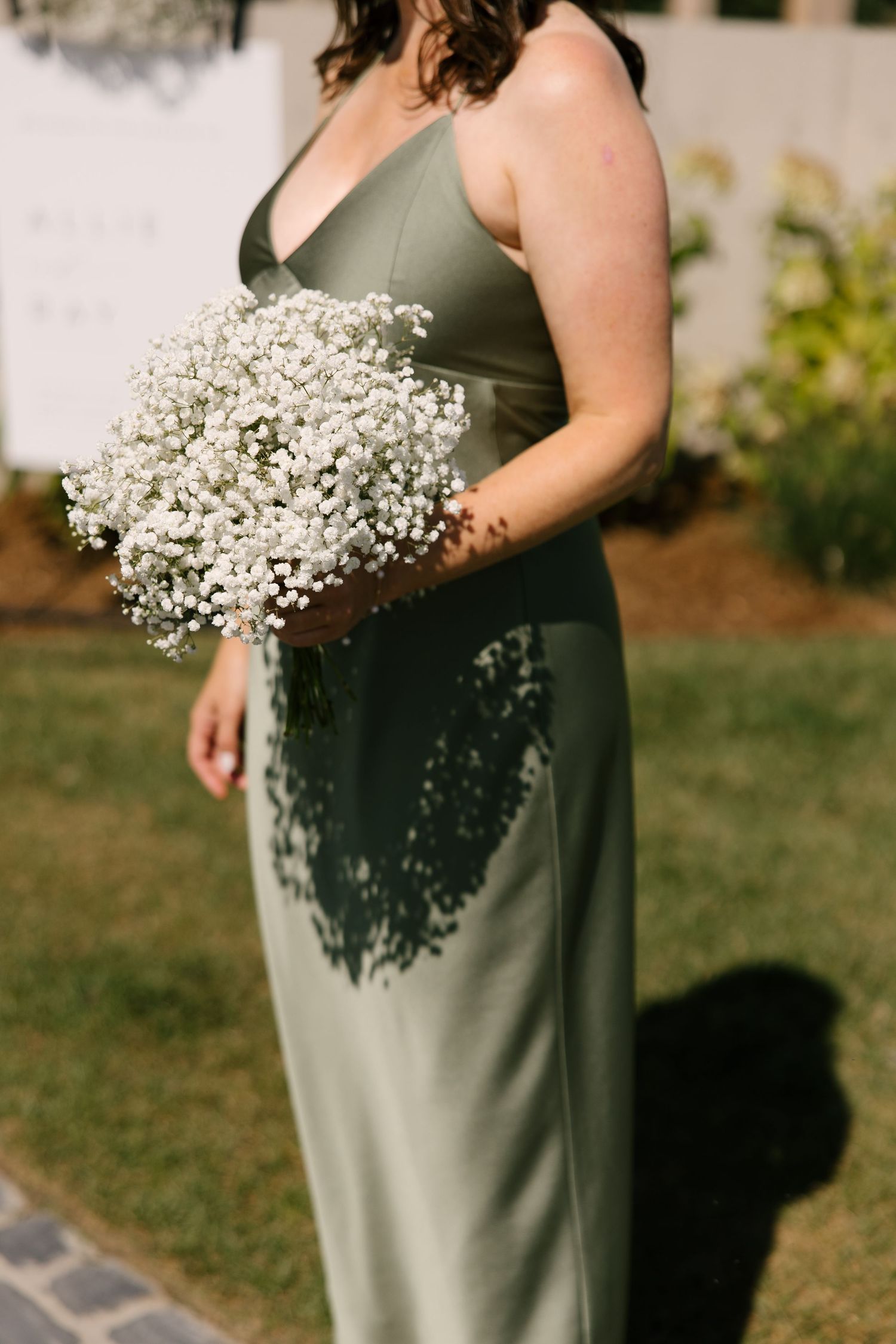 Bridesmaid in sage green gown holds lush white baby's breath bouquet in outdoor setting.