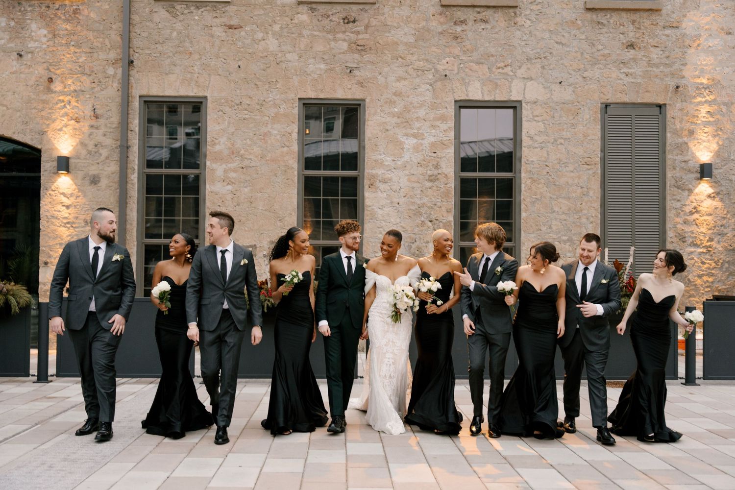 Wedding party in black attire walks with bride in white gown outside a rustic stone Tapestry Hall wedding venue.