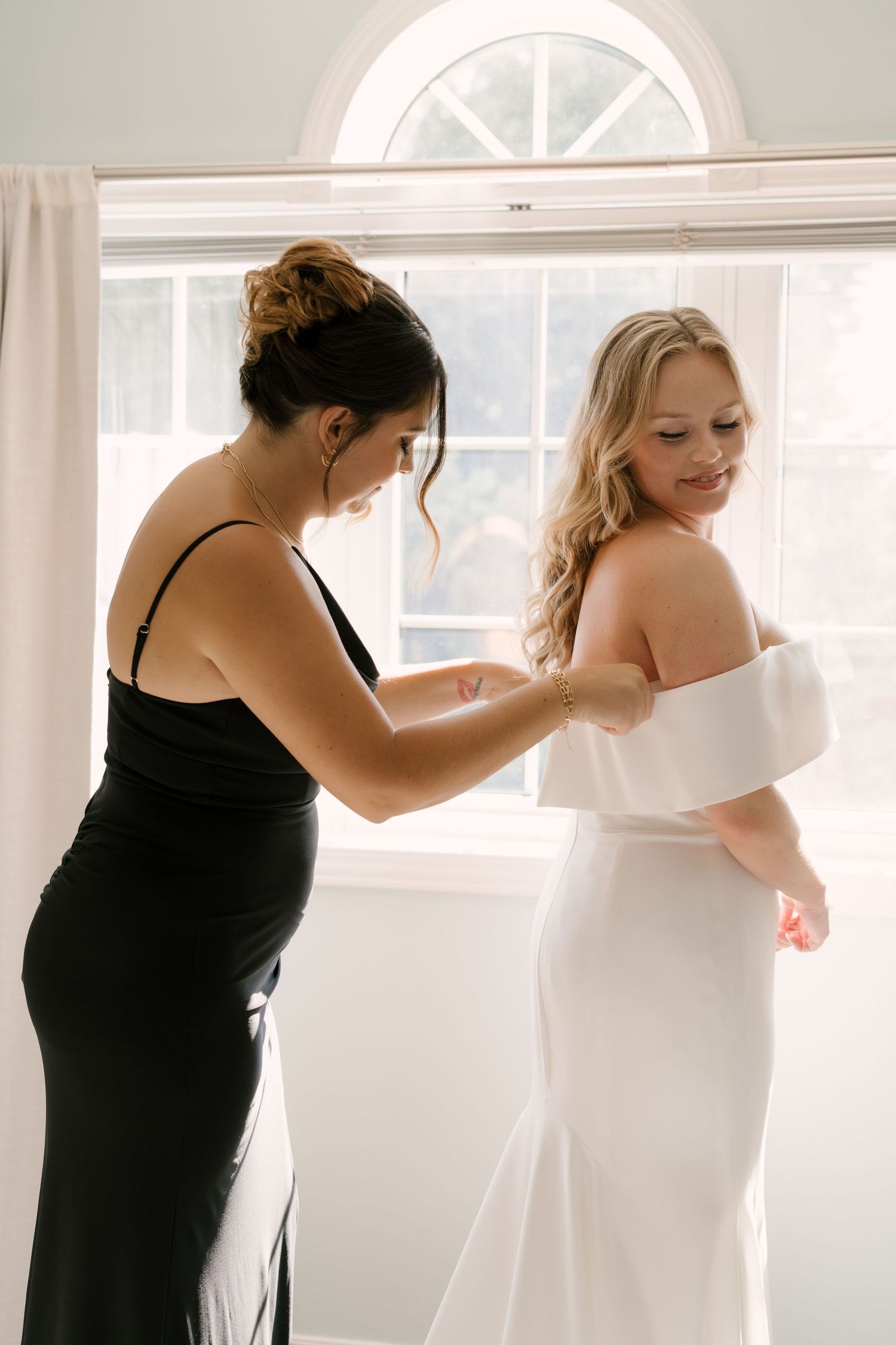 Bridesmaid in black dress helps blonde bride fasten her off-shoulder white wedding gown by window.