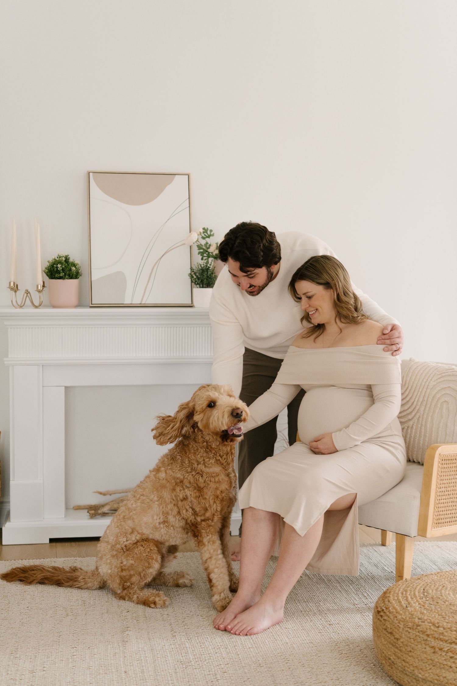 Couple with a golden doodle dog near a fireplace, pregnant woman seated cradling her bump while partner embraces her.