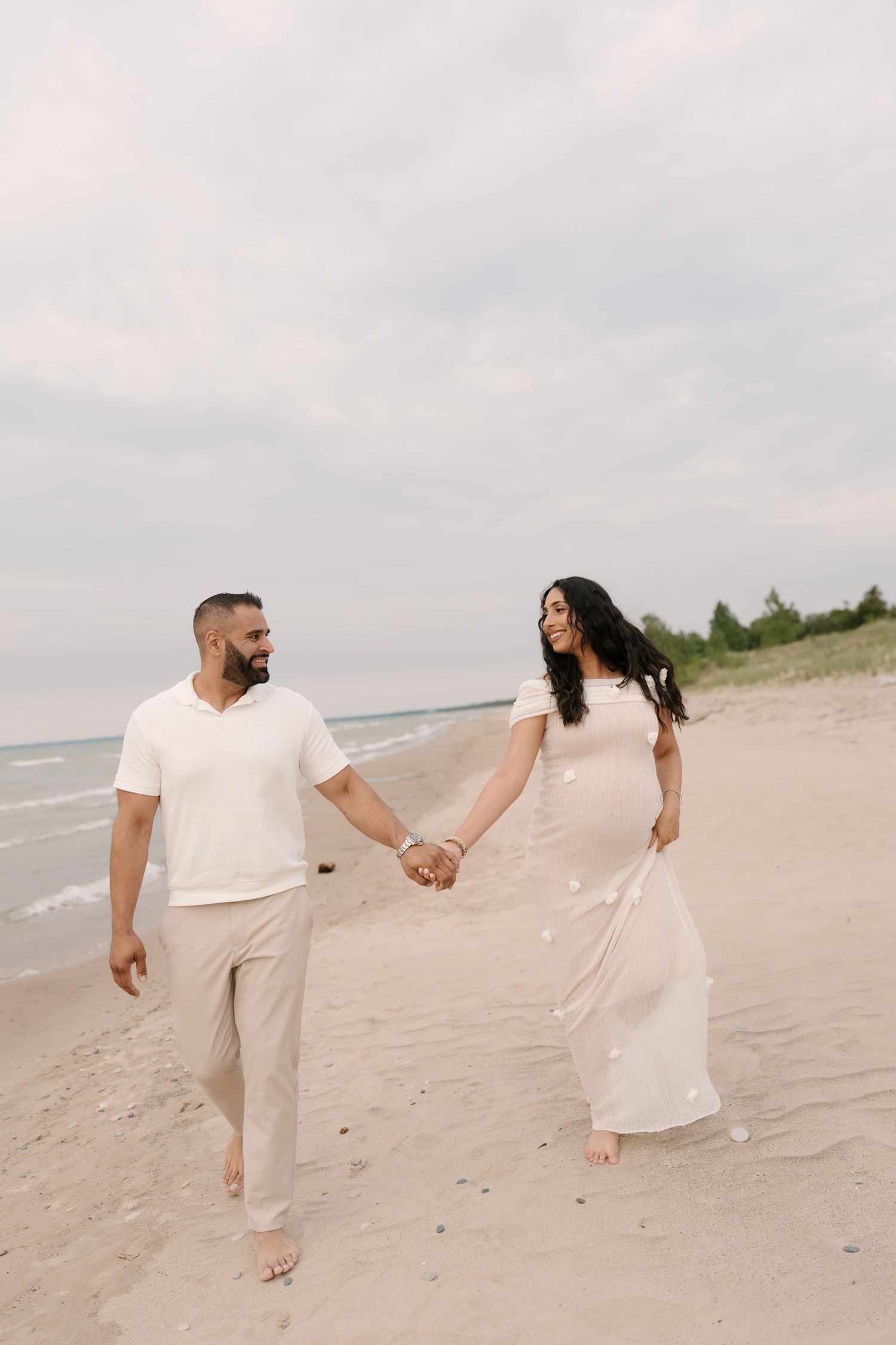 Smiling couple walks hand-in-hand along sandy beach, pregnant woman in flowing white dress with floral details.