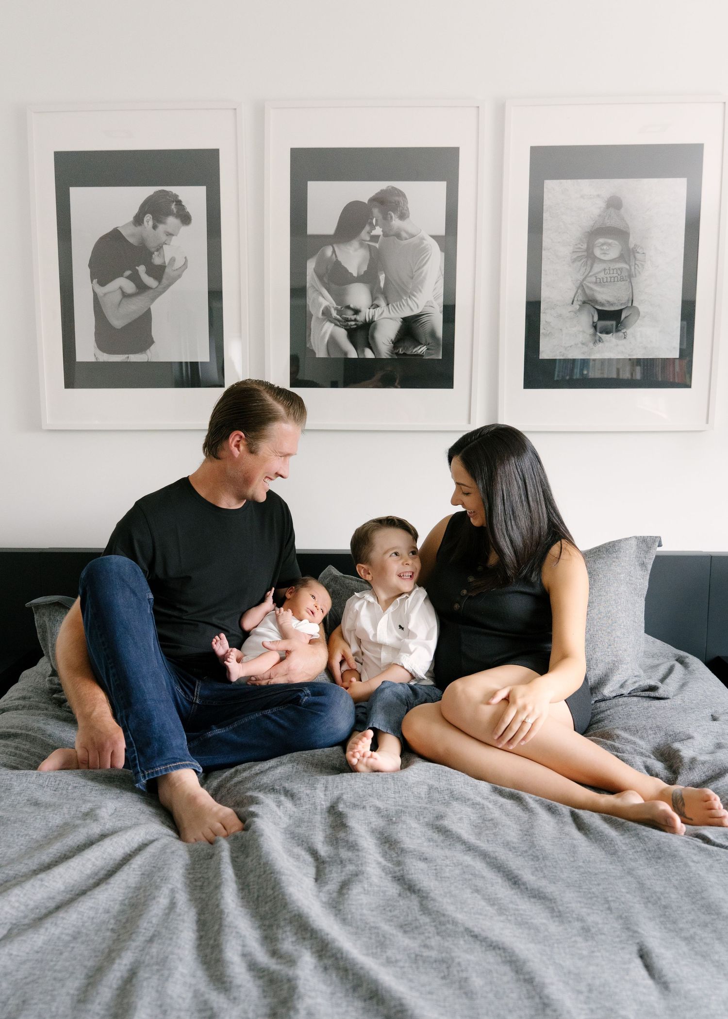 Happy family of four sitting on bed with black and white framed photos displayed on wall behind them.