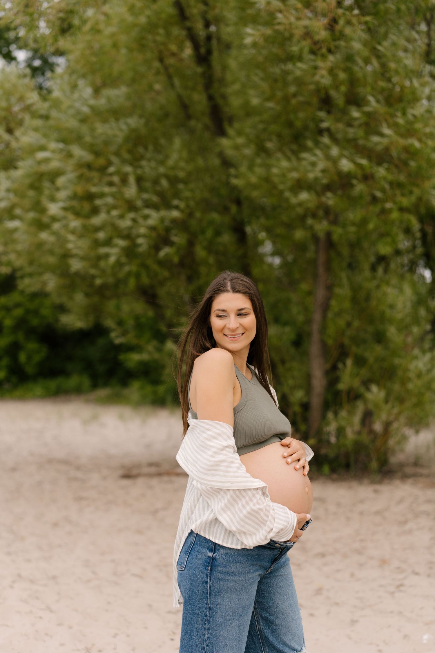 Smiling pregnant woman in olive crop top and open striped shirt cradles her baby bump on a sandy beach.