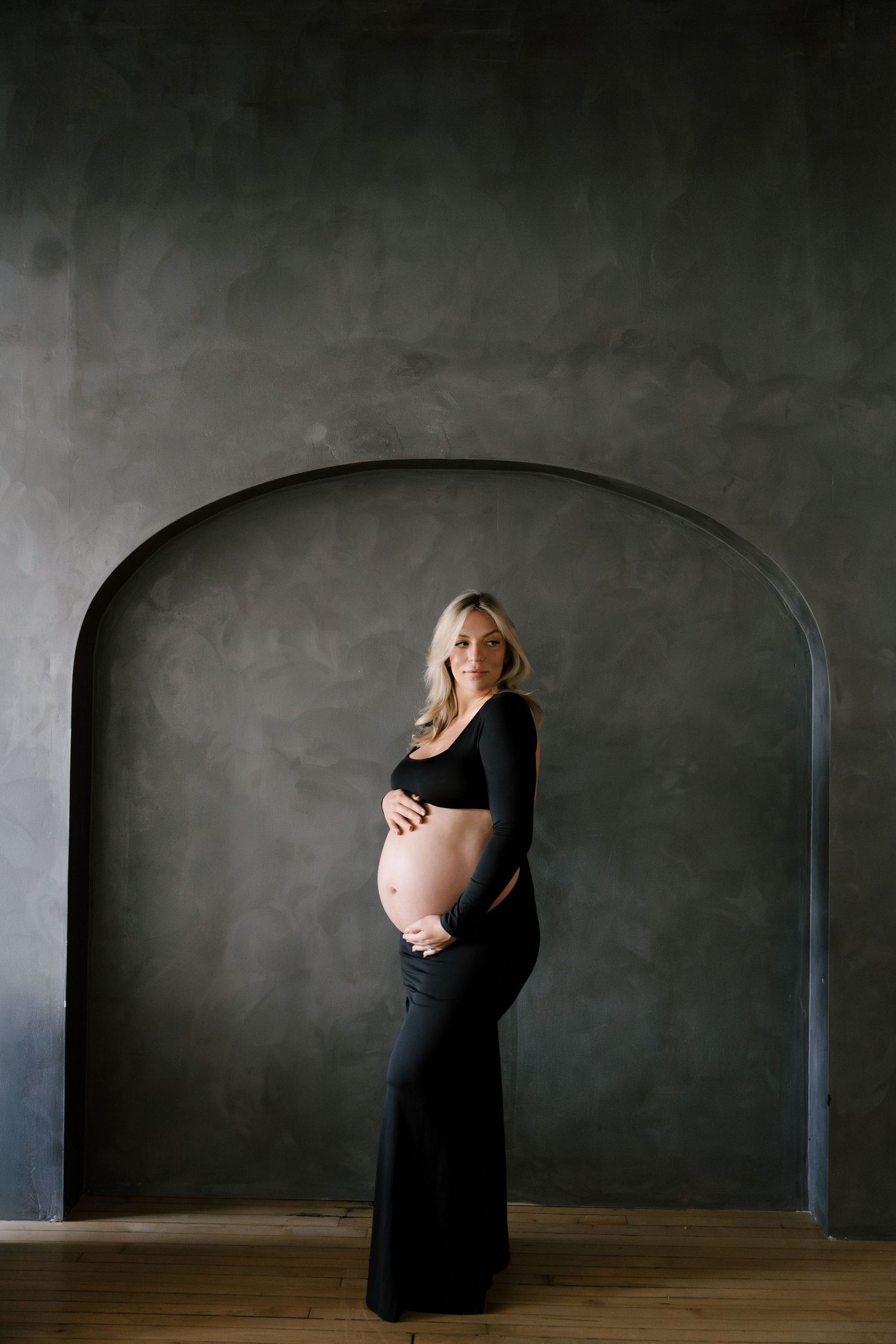 Pregnant blonde woman in black crop top and skirt holding belly, standing before arched gray wall.
