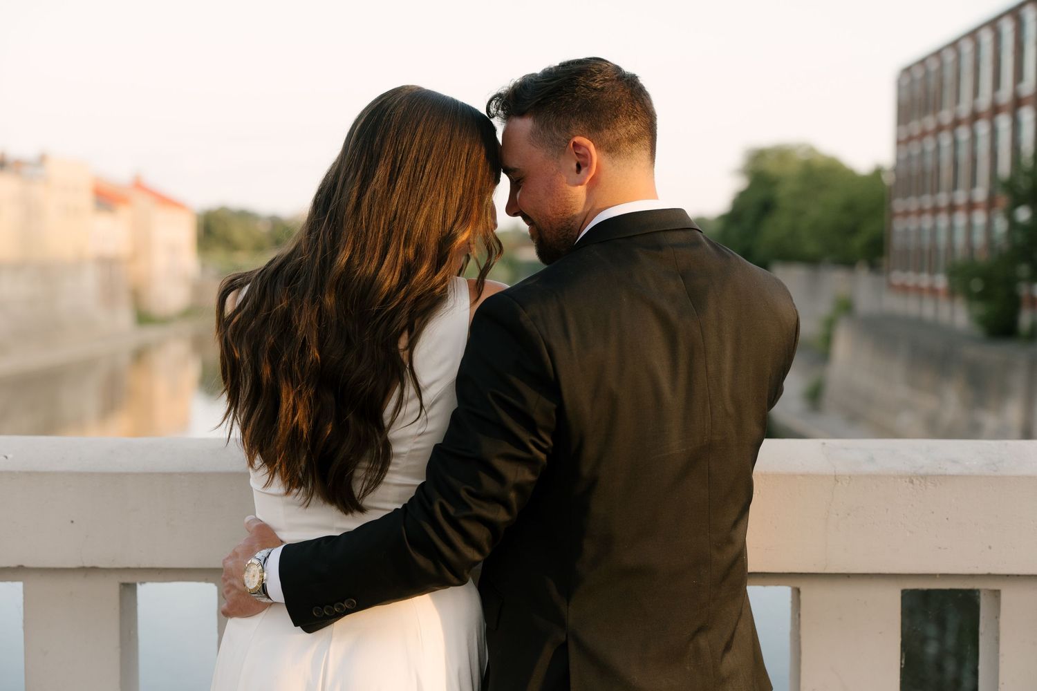 Couple embracing from behind on a bridge overlooking a scenic waterway at dusk.