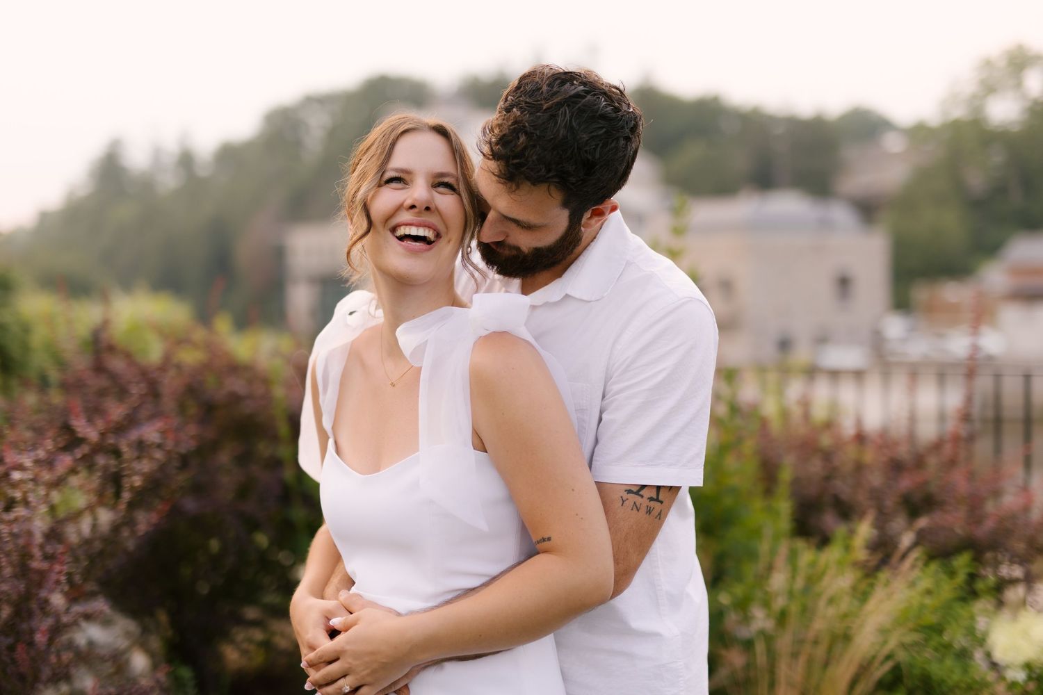 Joyful couple embracing outdoors with the woman laughing in a white dress against a garden backdrop.