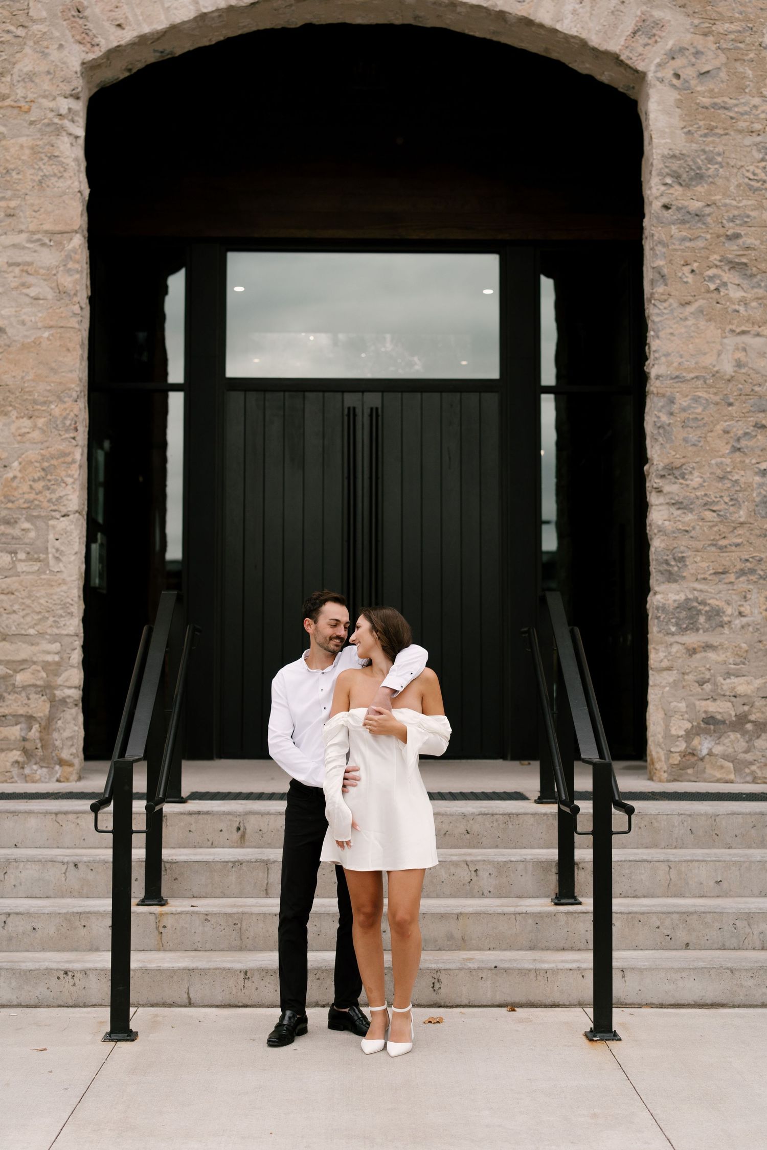 Engaged couple embracing on stone building steps, woman wearing white mini dress and heels.