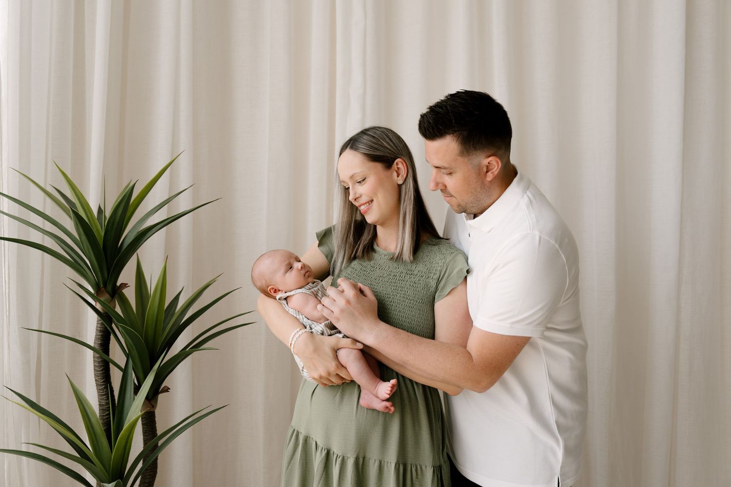 A couple stands by a green plant holding their newborn baby, mother in a sage green dress.