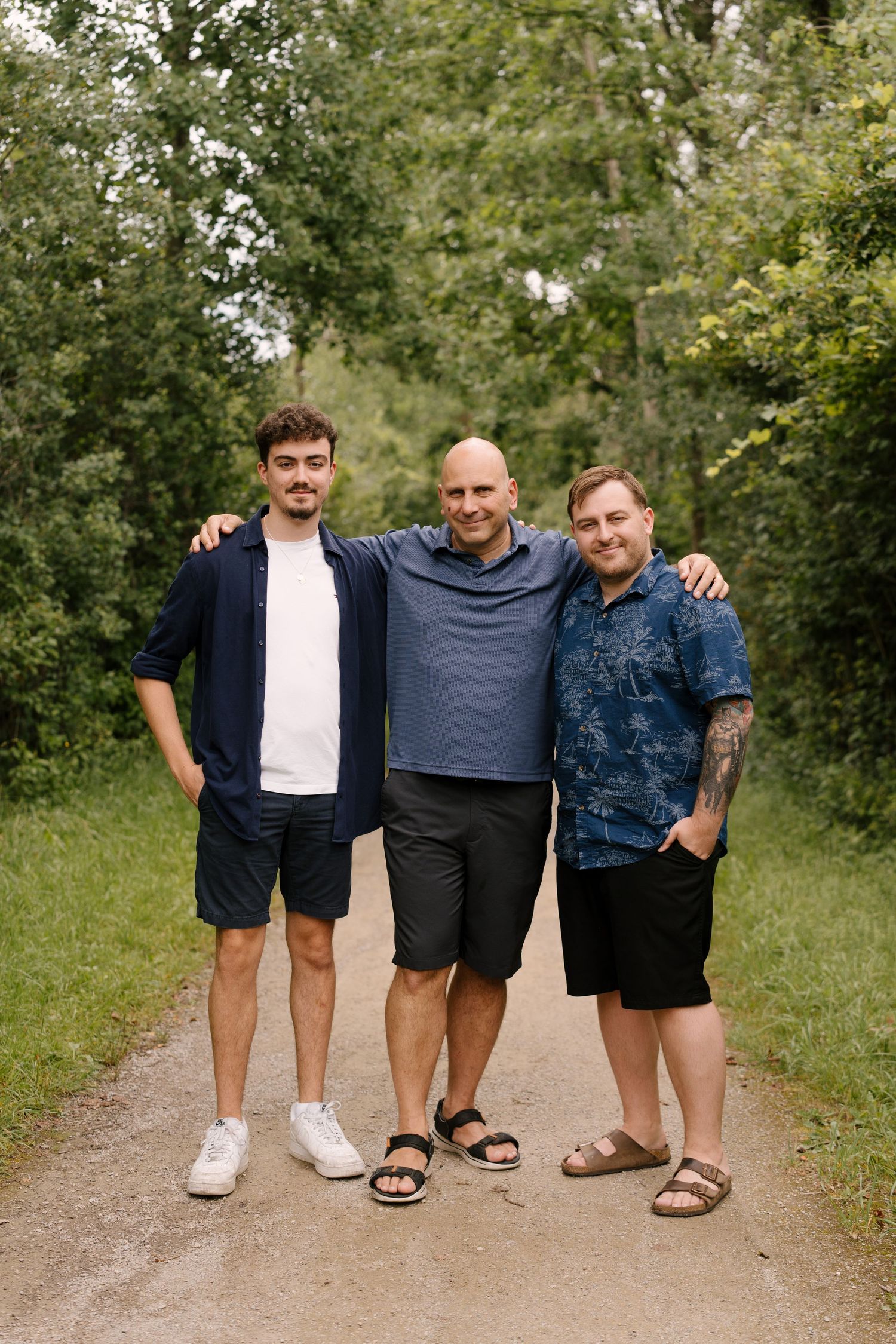 Three men smiling and posing together on a tree-lined path outdoors on a sunny day.