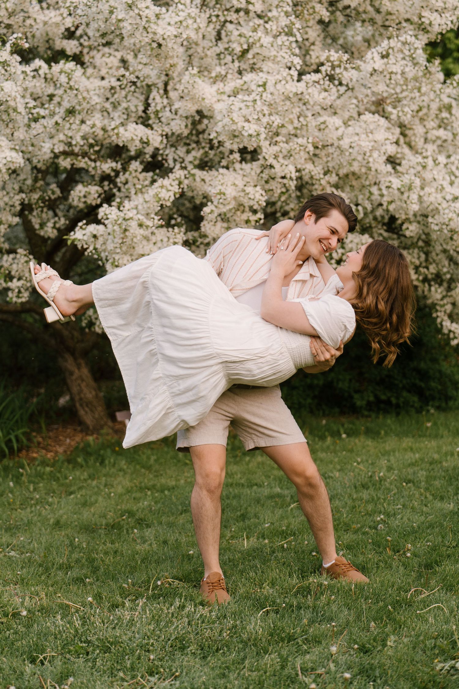 Man joyfully lifting woman in his arms while sharing a romantic moment in front of blooming white flowers.
