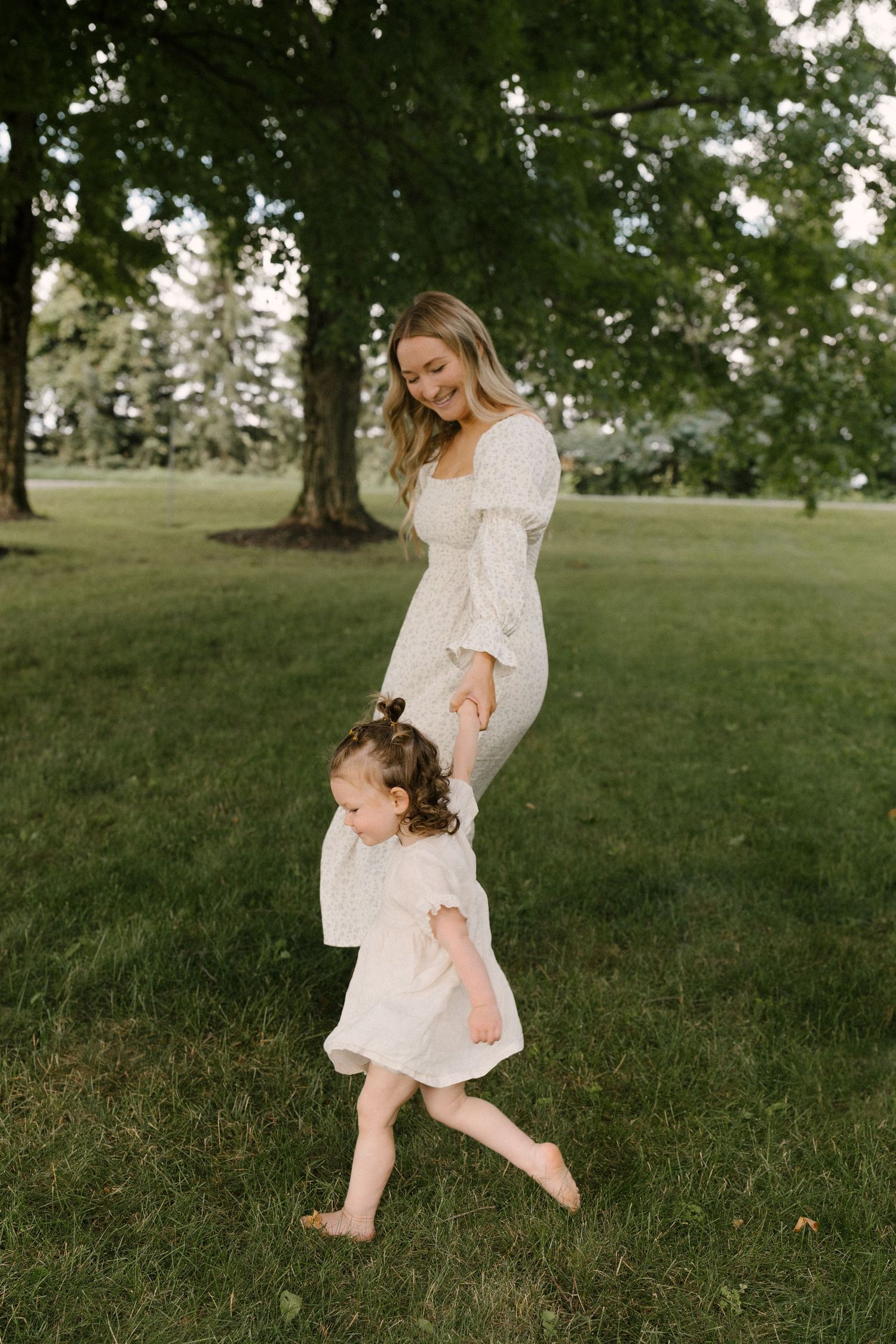 A smiling mother in a white dress holds her toddler daughter's hand while walking barefoot on green grass.